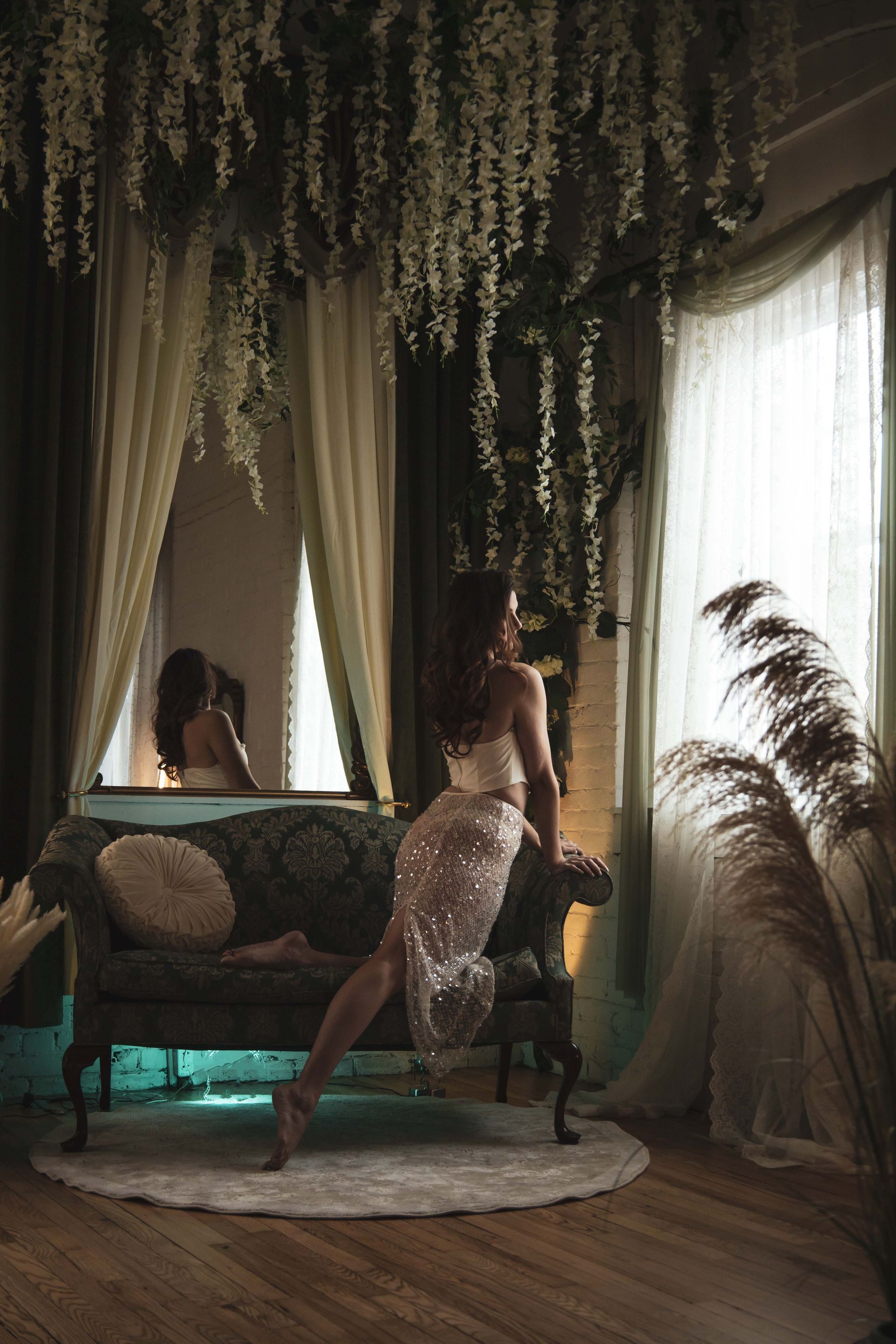 Woman in sparkly dress leans on an ornate sofa, looking out a window. Soft lighting and hanging greenery create an ethereal atmosphere.
