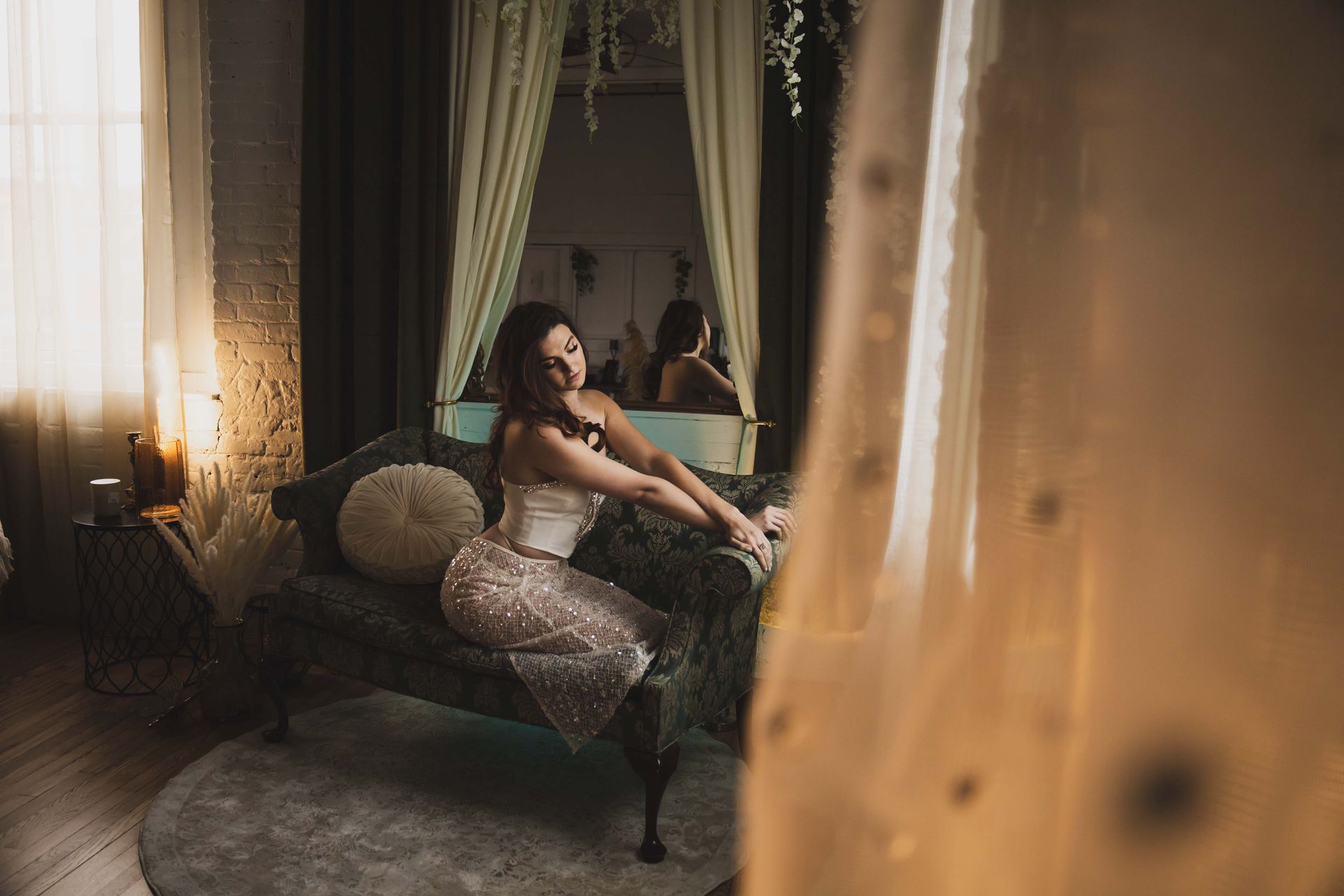 Woman in a sparkly skirt and corset poses on a green velvet couch in a softly lit room.