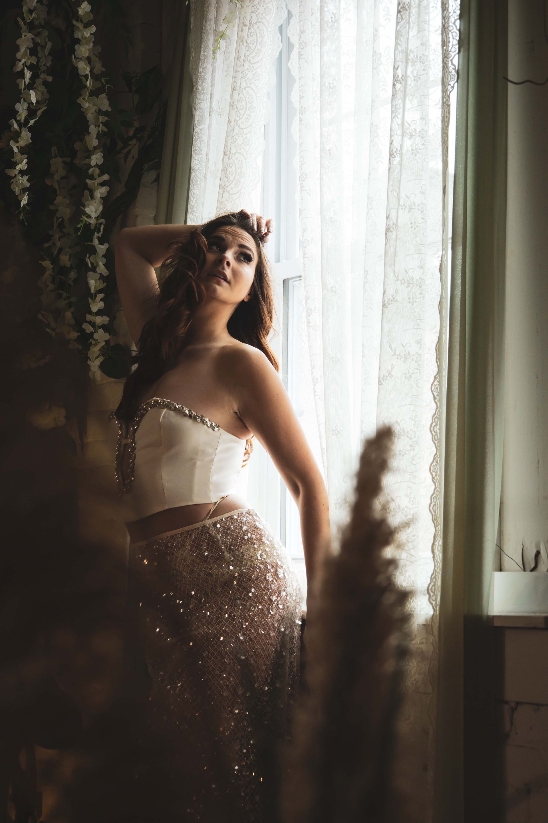 Woman in sequined skirt and white bustier poses by a lace-curtained window, arm raised.