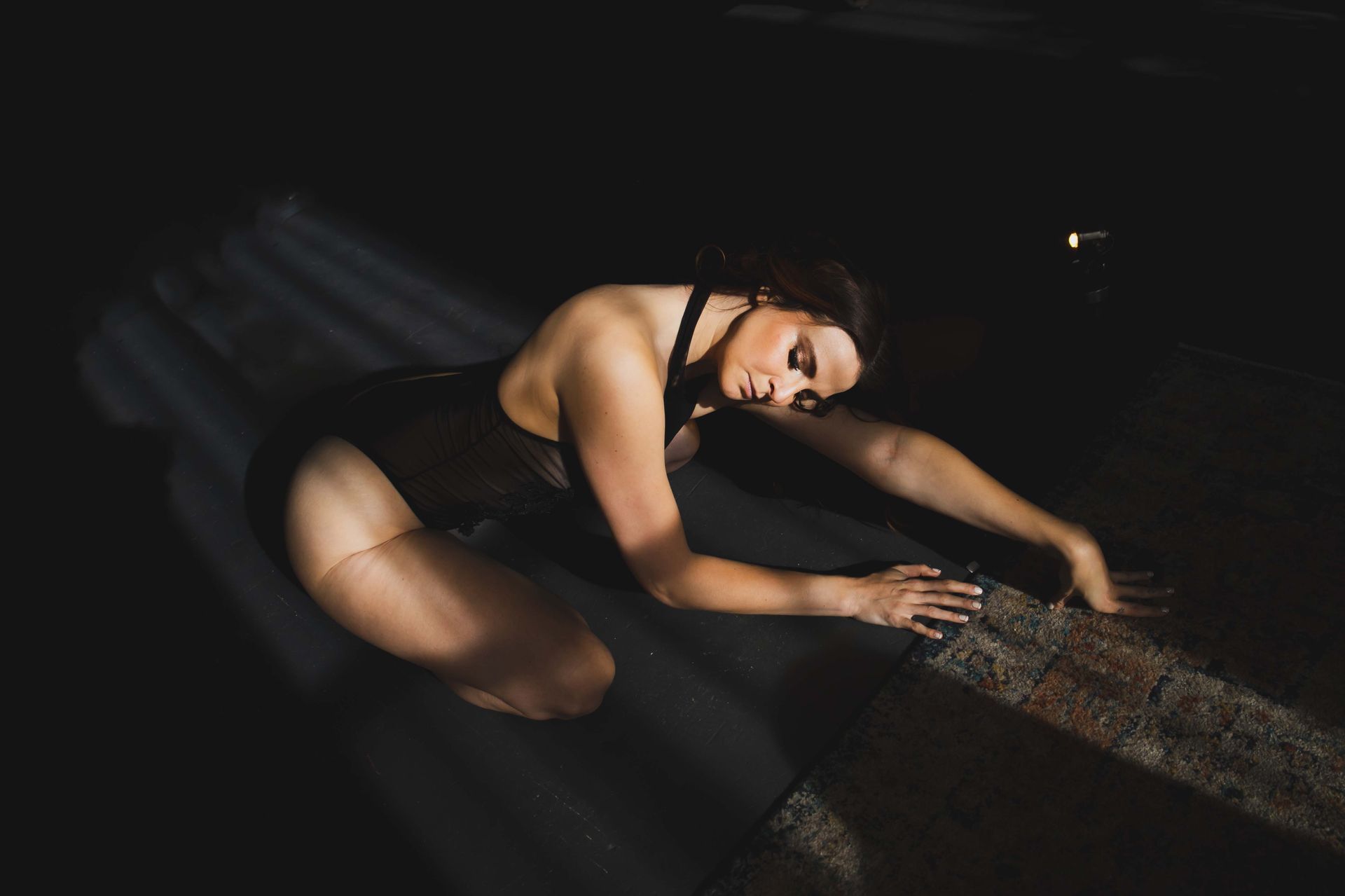Woman in a black leotard kneels, arms outstretched, in a dark space with a beam of light.