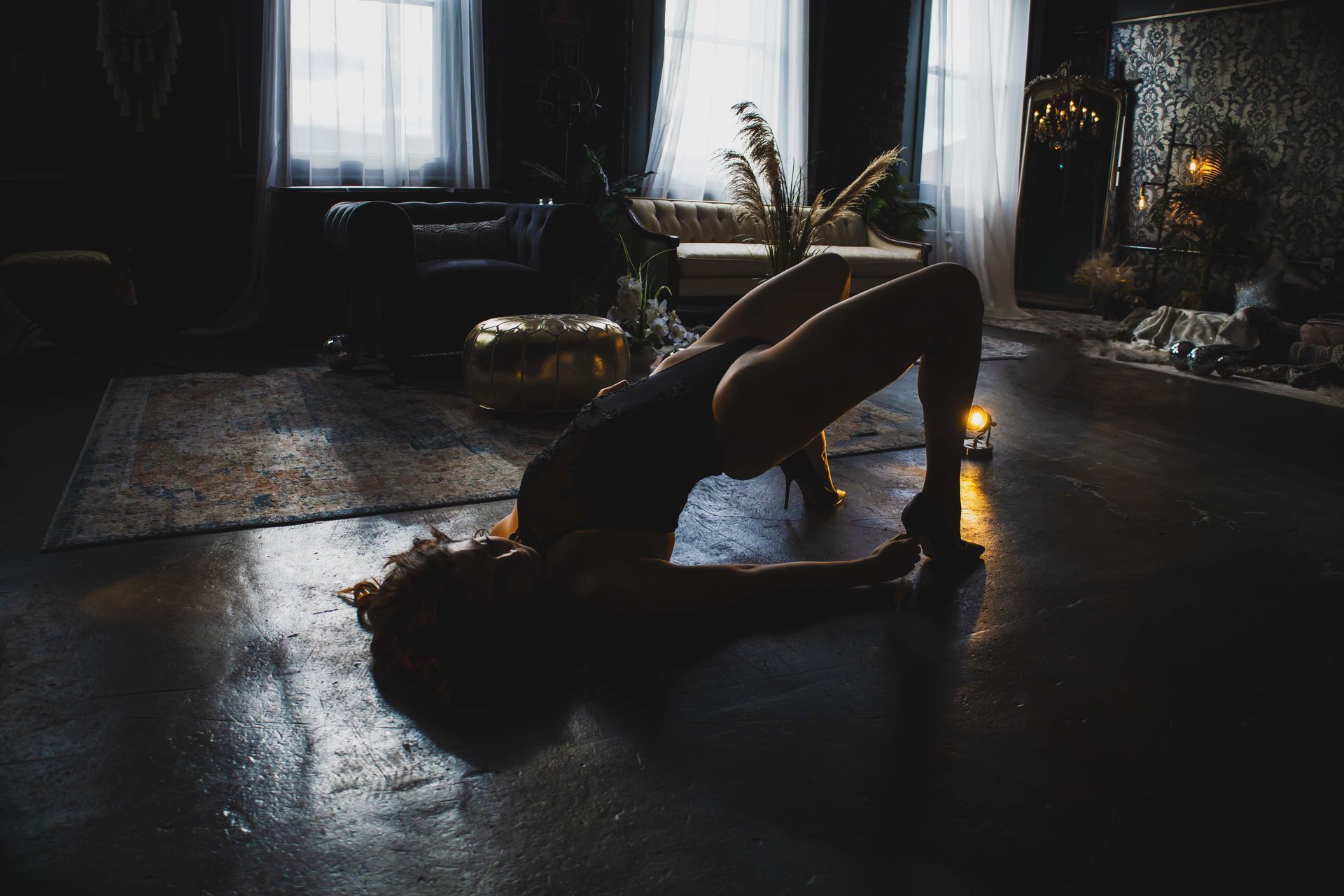 Woman in a dark room doing a bridge pose, floor reflects light, near a rug and windows.