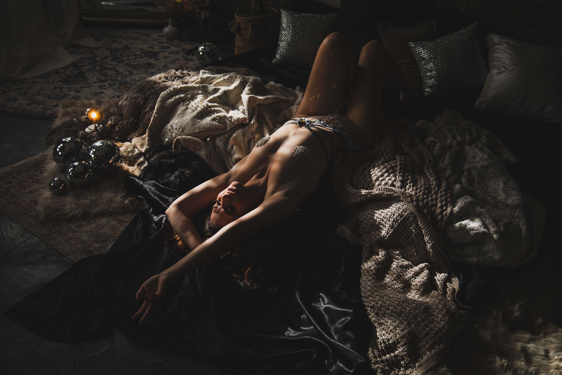 Woman reclining on a bed with textured blankets, illuminated by warm light. She is looking up with one arm outstretched.