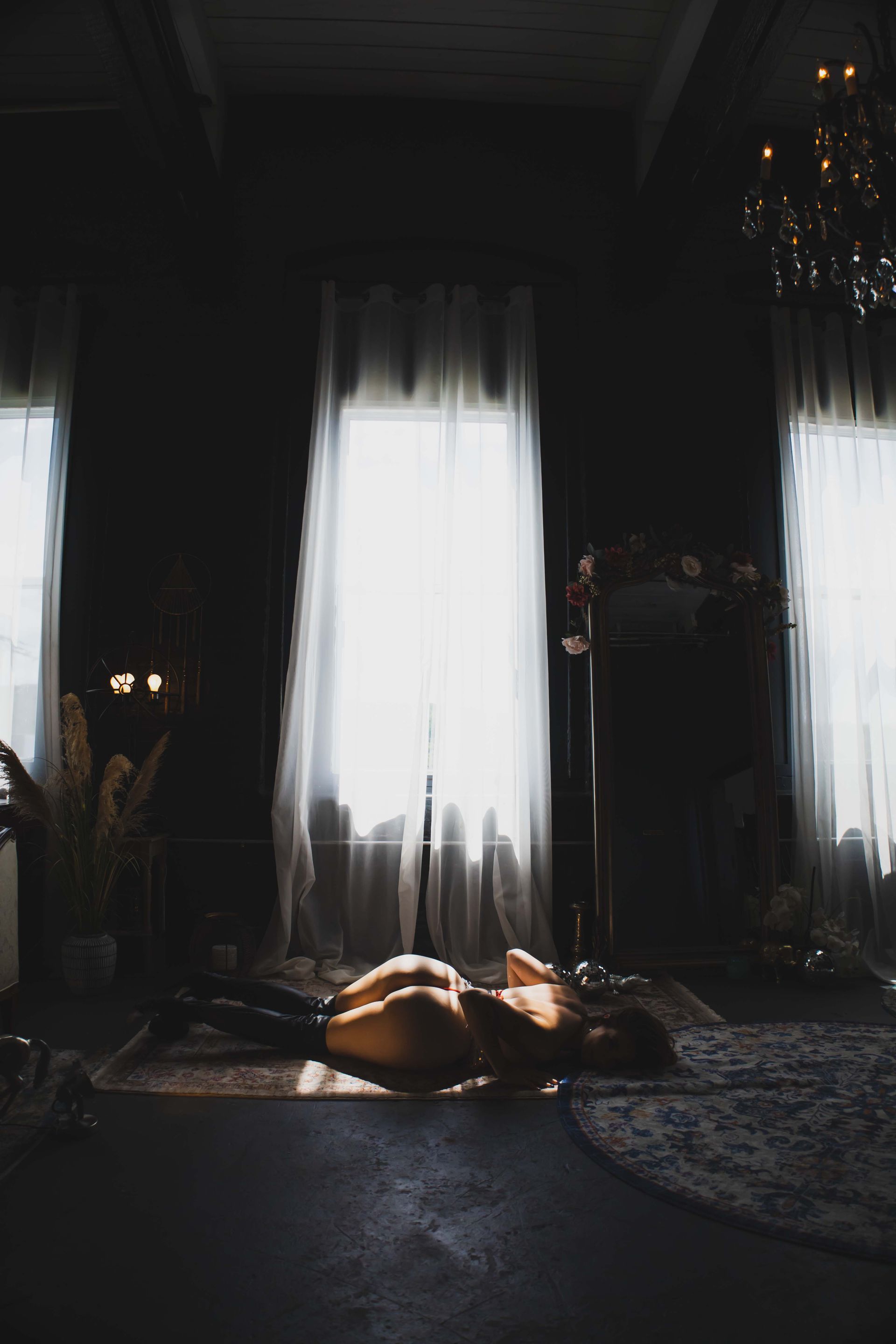 A person lies on a rug in a dimly lit room, bathed in light from a window.  A chandelier and dark furnishings are visible.