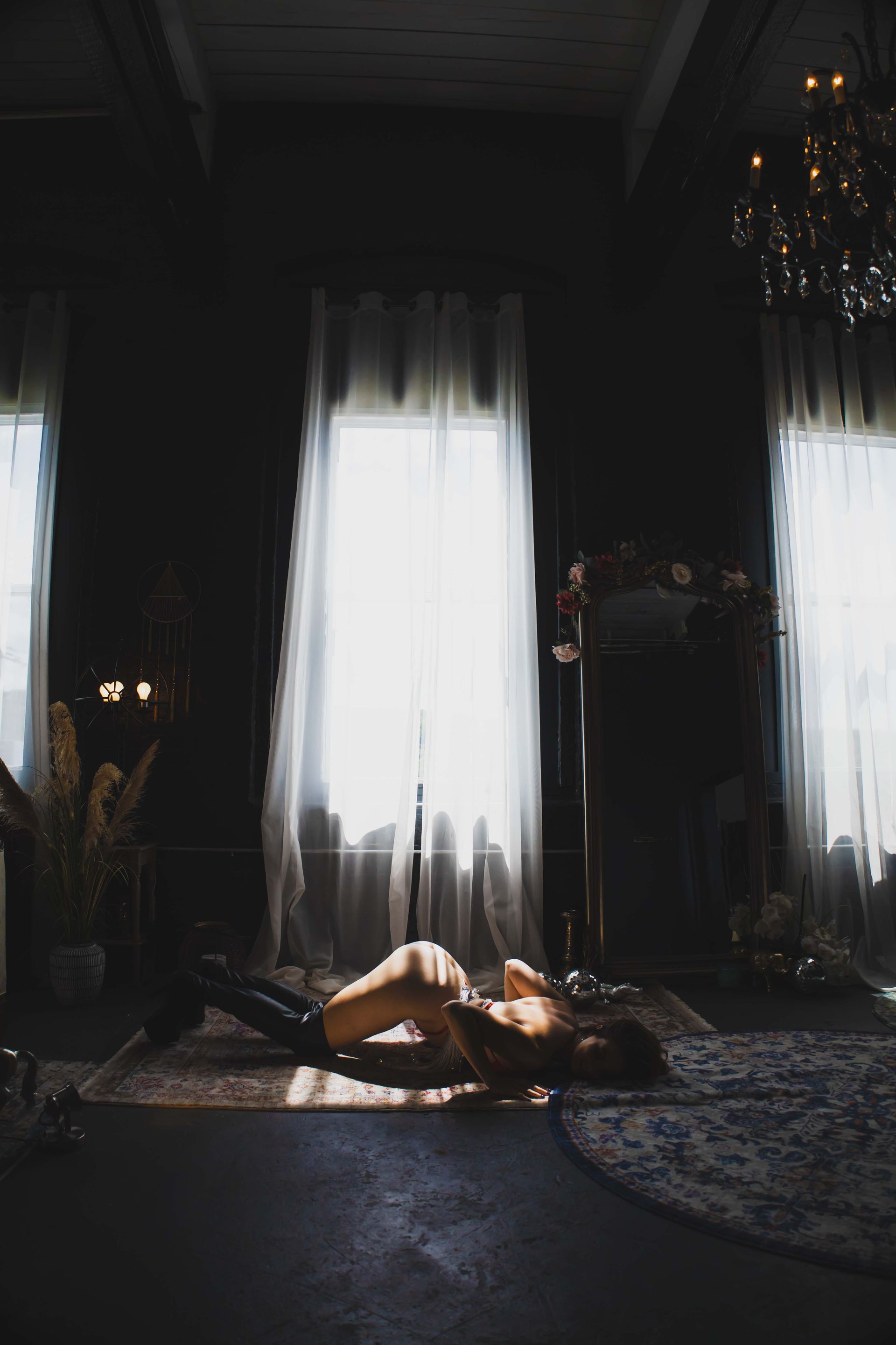 Woman doing yoga on a patterned rug near a bright window, in a dark room. A chandelier hangs above.