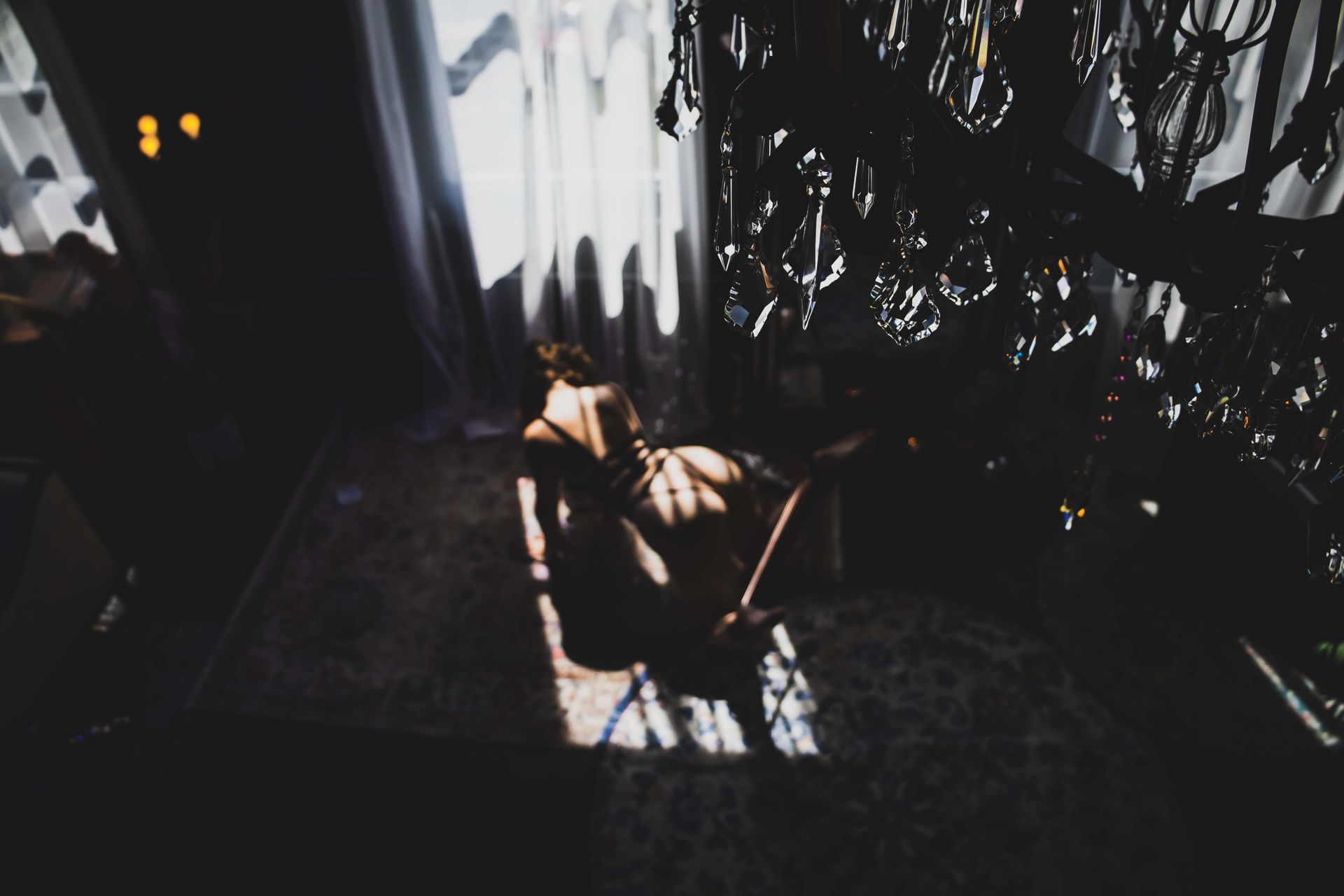 Woman in lingerie sits on rug near a window with sunlight shining through. Chandelier is visible.