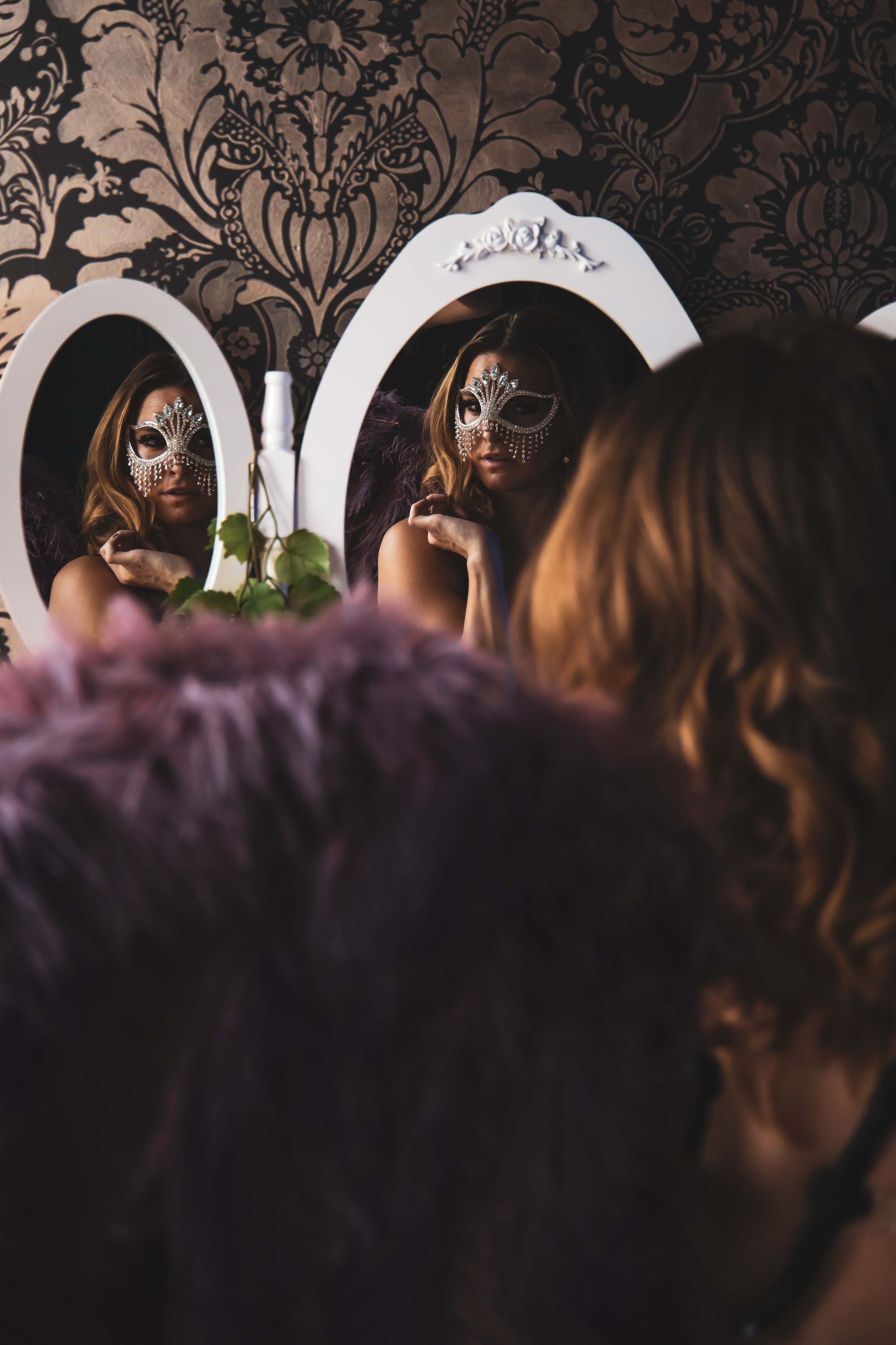 Woman in a mask reflected in two mirrors, wearing a fur coat. Wallpapered room in shades of brown and purple.
