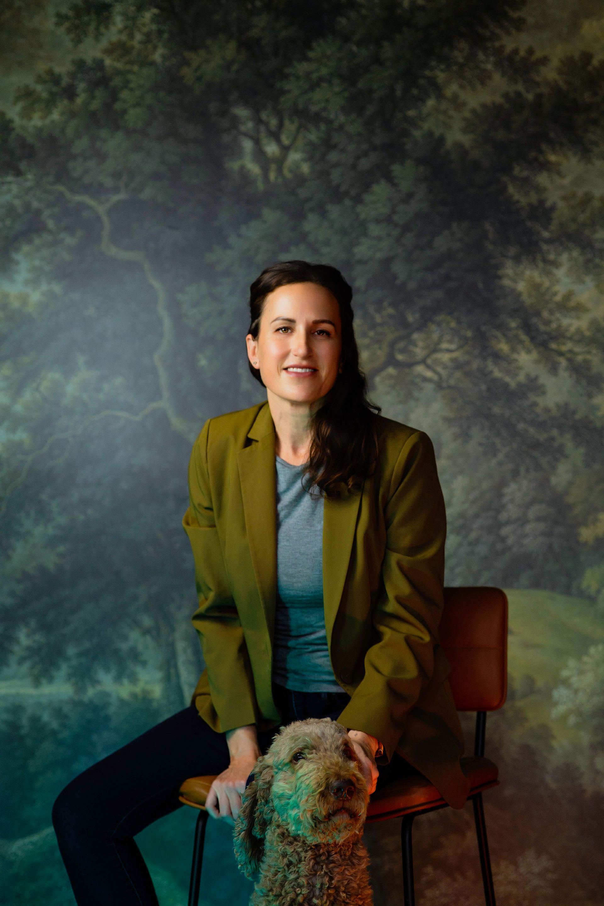 Portrait of a seated person in a green jacket with a dog, against a painted forest backdrop