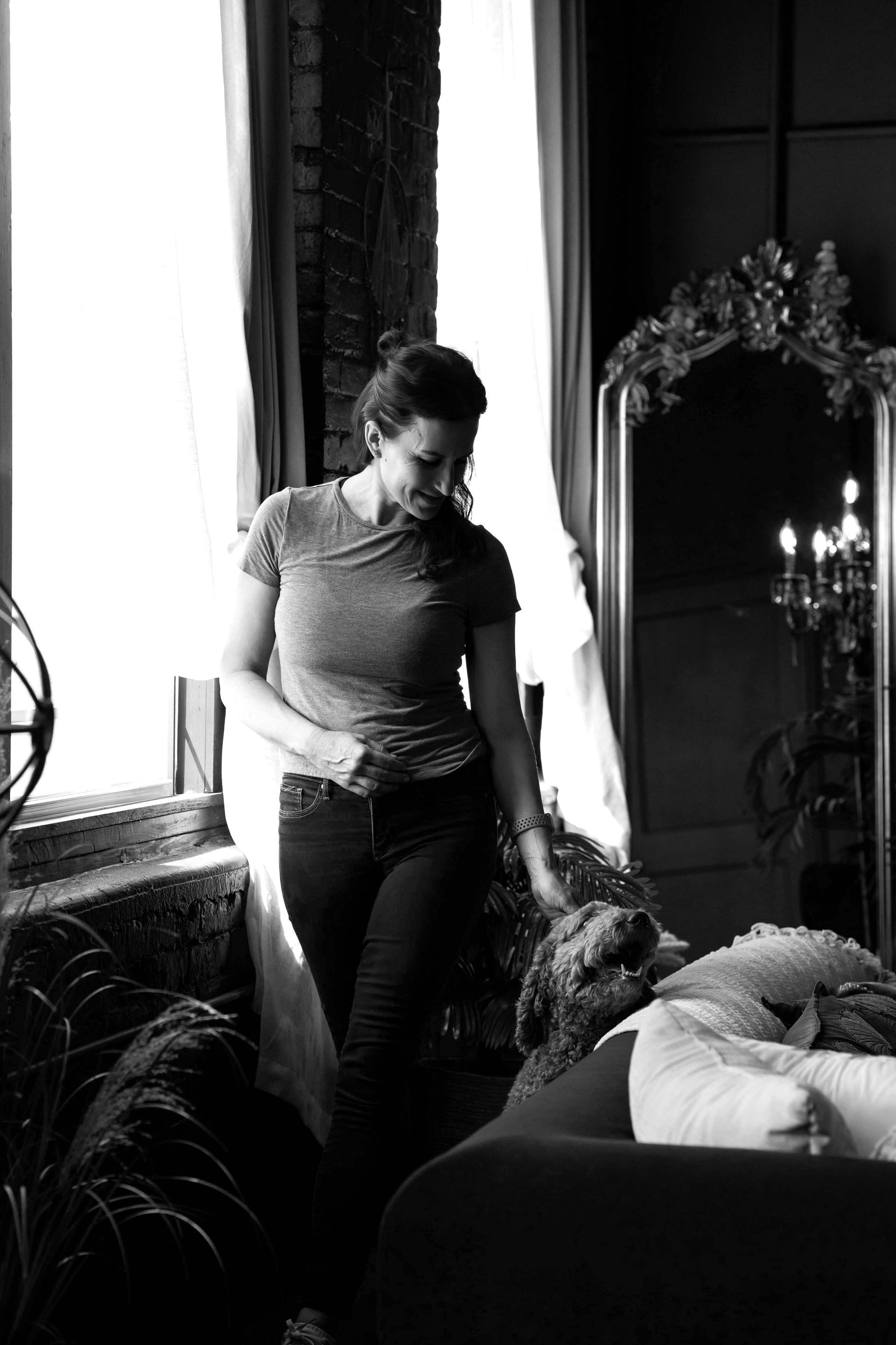 Woman standing beside a couch in a dimly lit room, looking down at a resting dog.