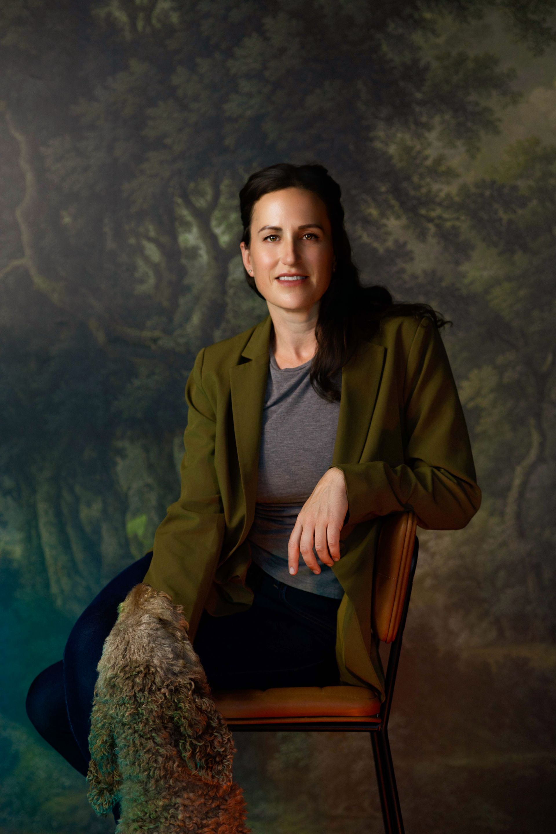 Person seated on a chair in a studio portrait, wearing an olive jacket and smiling against a painted backdrop