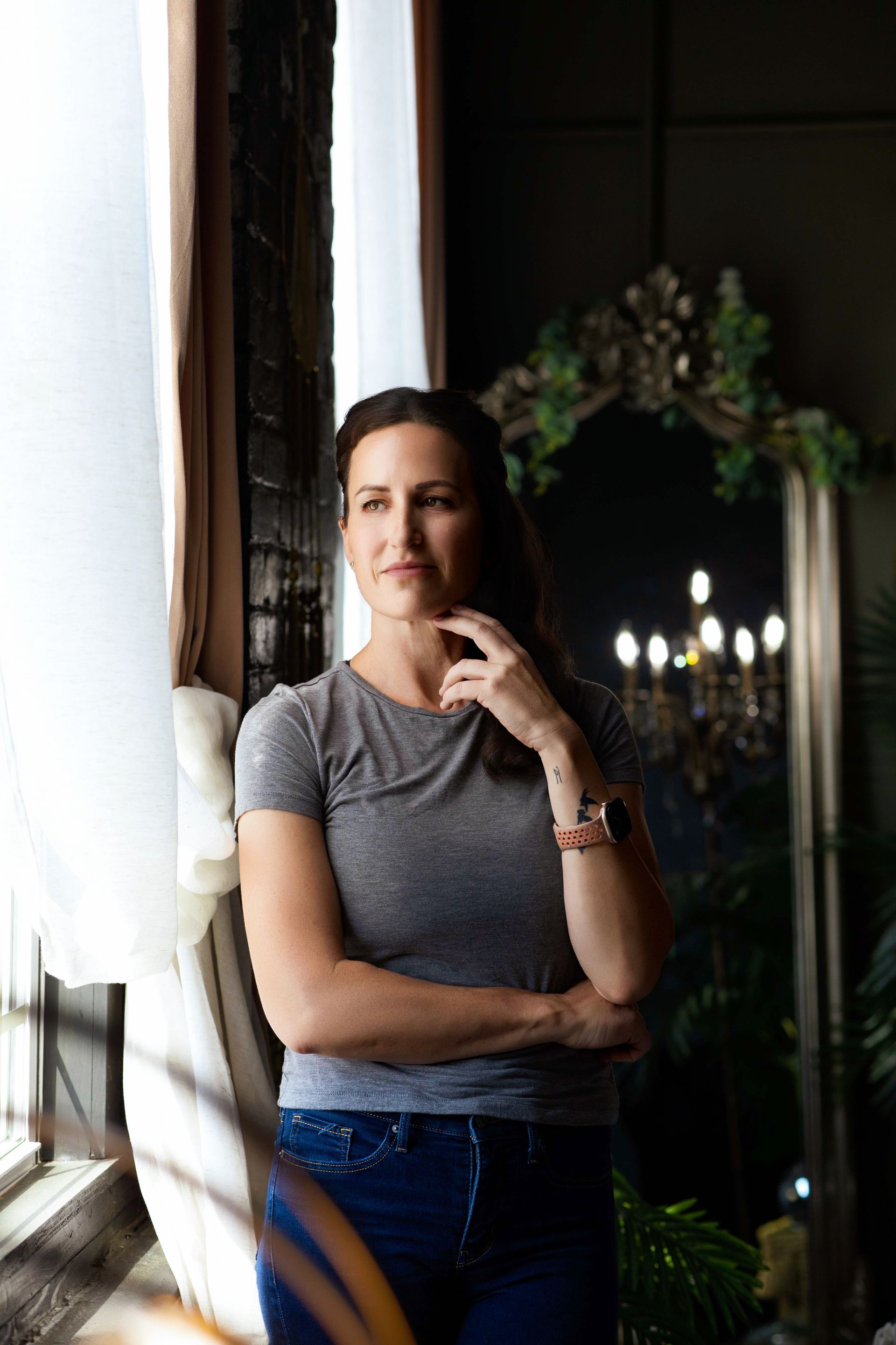 Woman standing by a window in a dim room, wearing a gray shirt and jeans, with flowers in the background