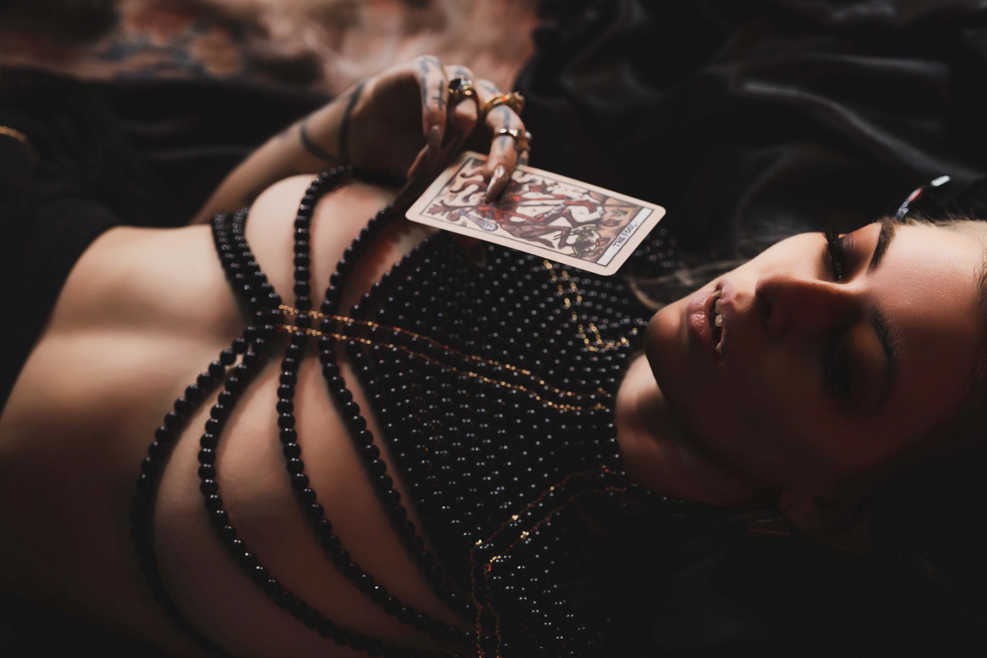 A person wearing a beaded harness holds up a tarot card while reclining. Moody lighting, dark colors, and jewelry.