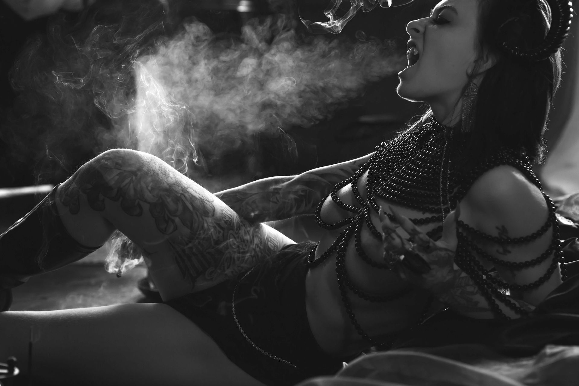 Woman with tattoos and dark attire exhales mist, horns, open mouth. Black and white setting.