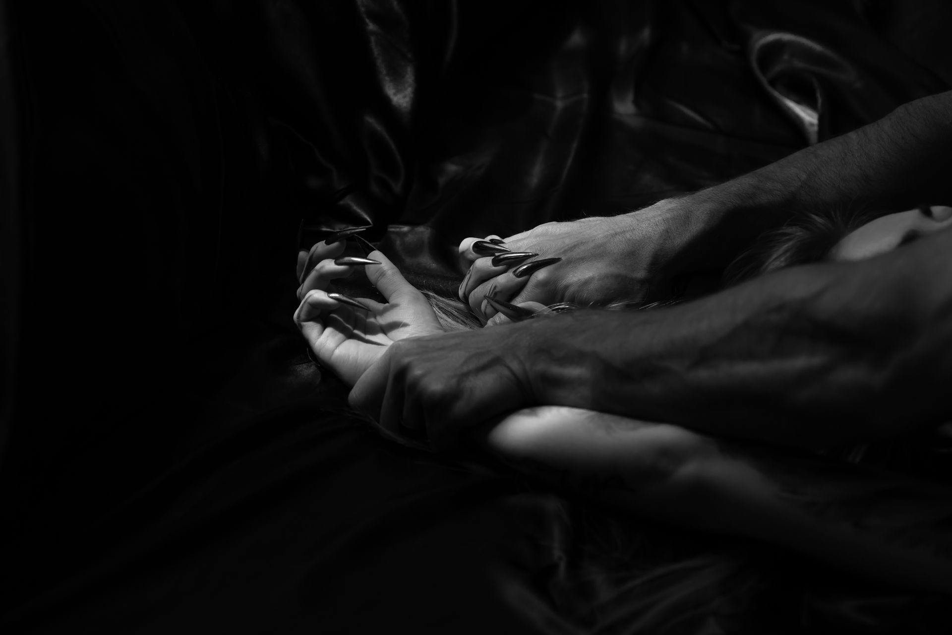 Hands gripping a person’s face and wrist against a dark, silky background, in black and white.