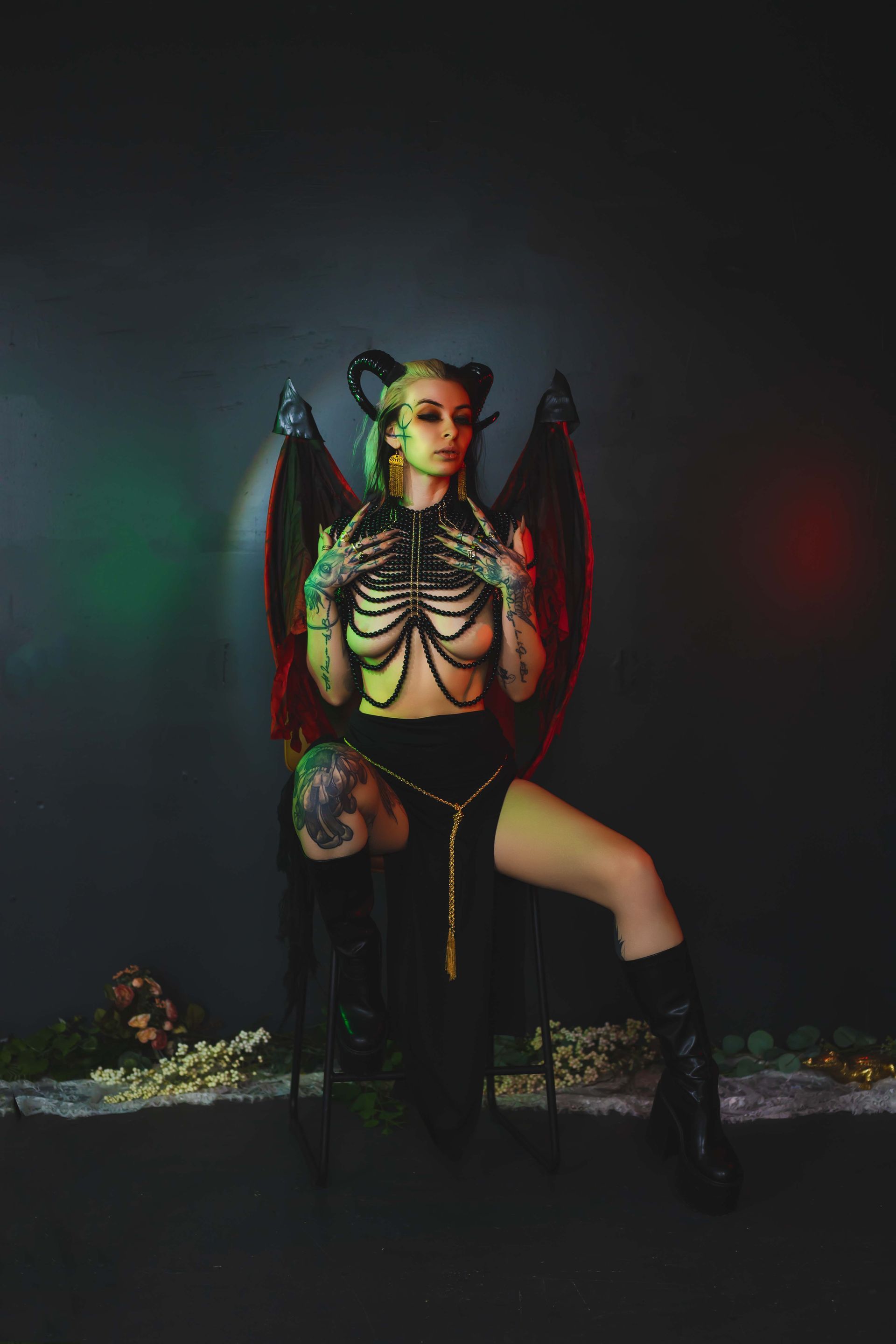 Woman with horns and wings in devil-themed costume, posed on a chair against a dark backdrop with green and red lighting.