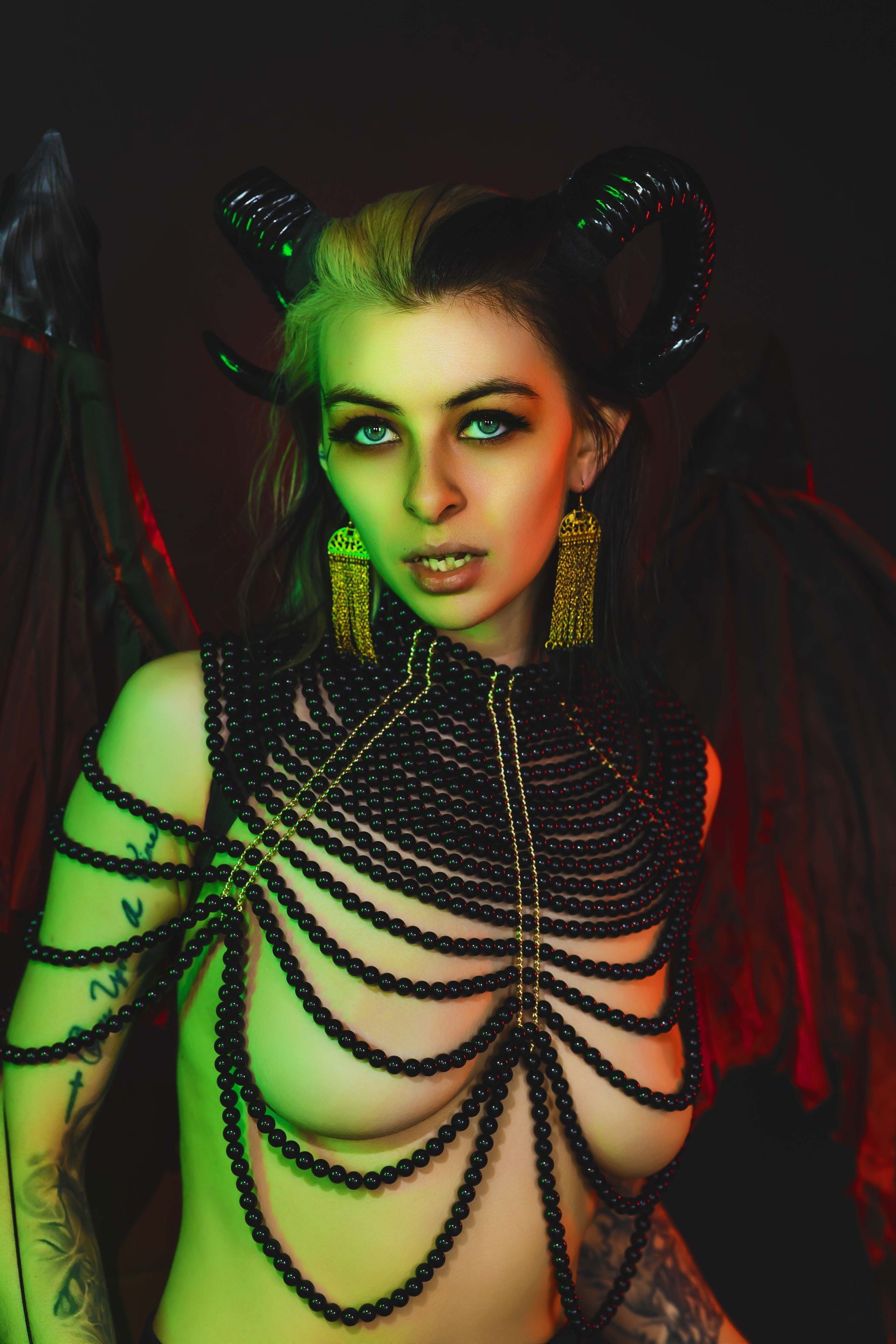 Woman in demon-like costume with horns, wings, and beaded body jewelry. Green and red lighting, looking directly at the viewer.