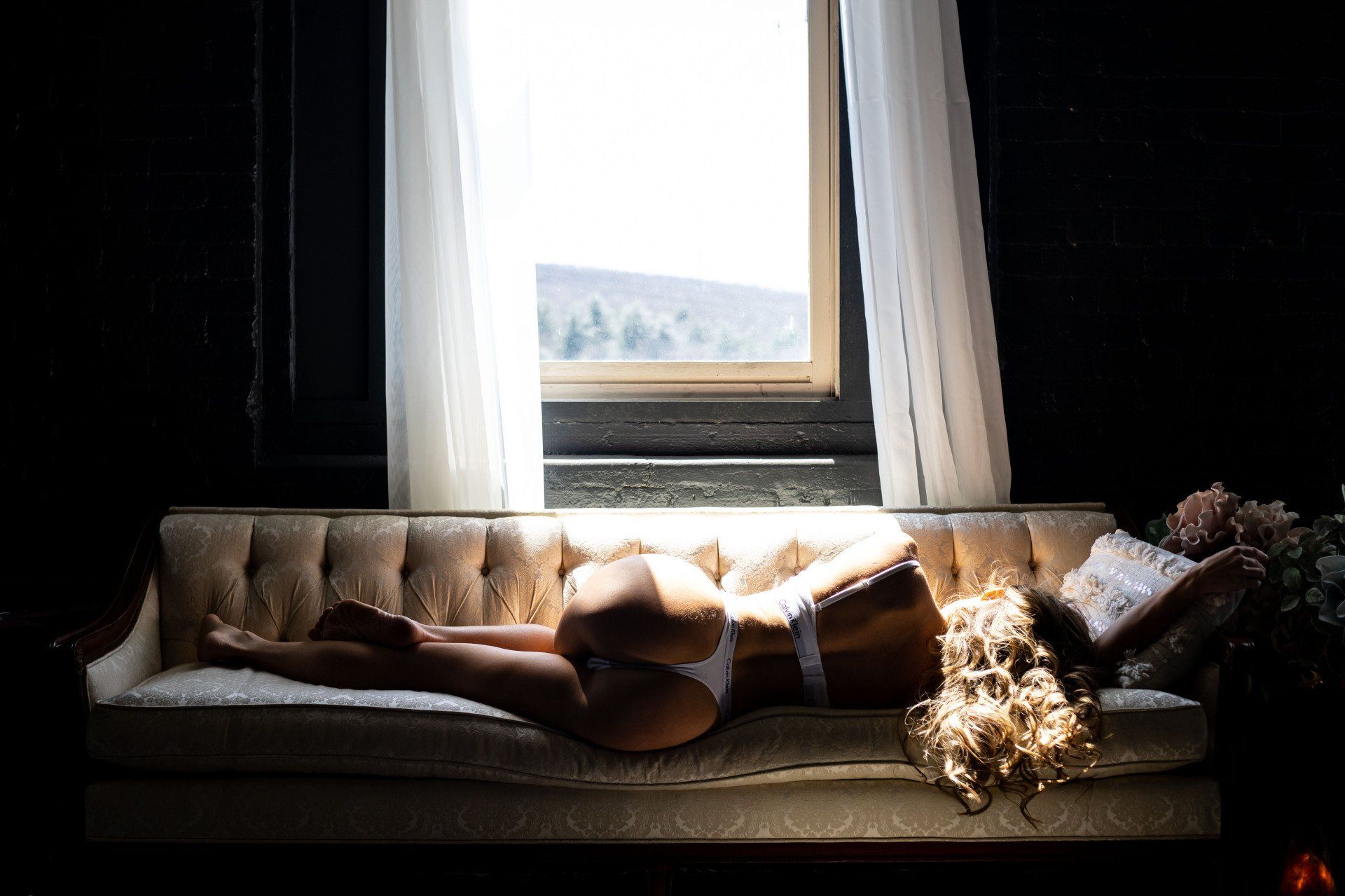 Woman in white lingerie reclines on a cream sofa, back to the camera, near a window. Sunlight illuminates the scene.