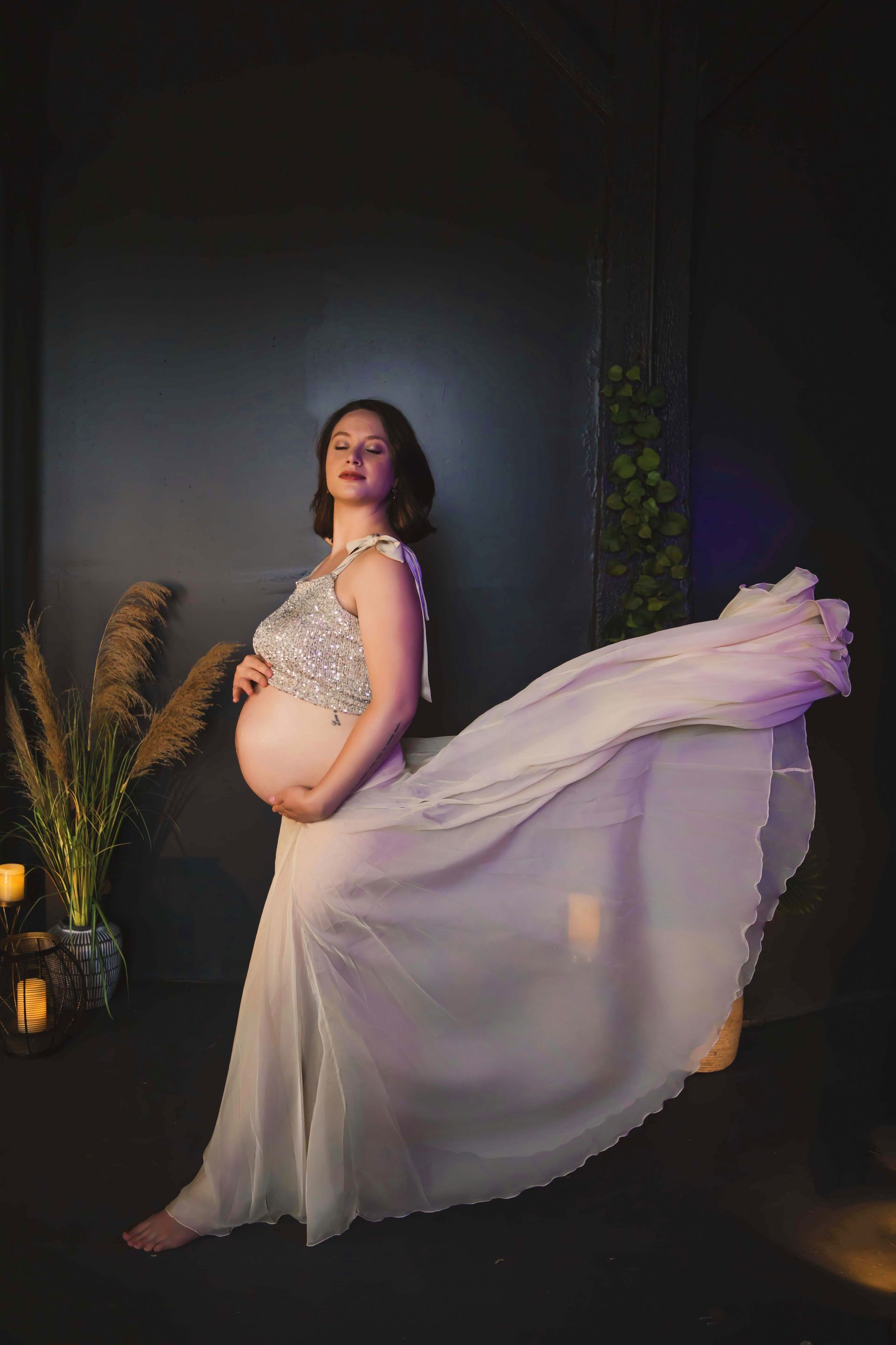 Pregnant woman in a sparkling top and flowing white dress, hand on her belly, in a dark studio setting with tall grass.