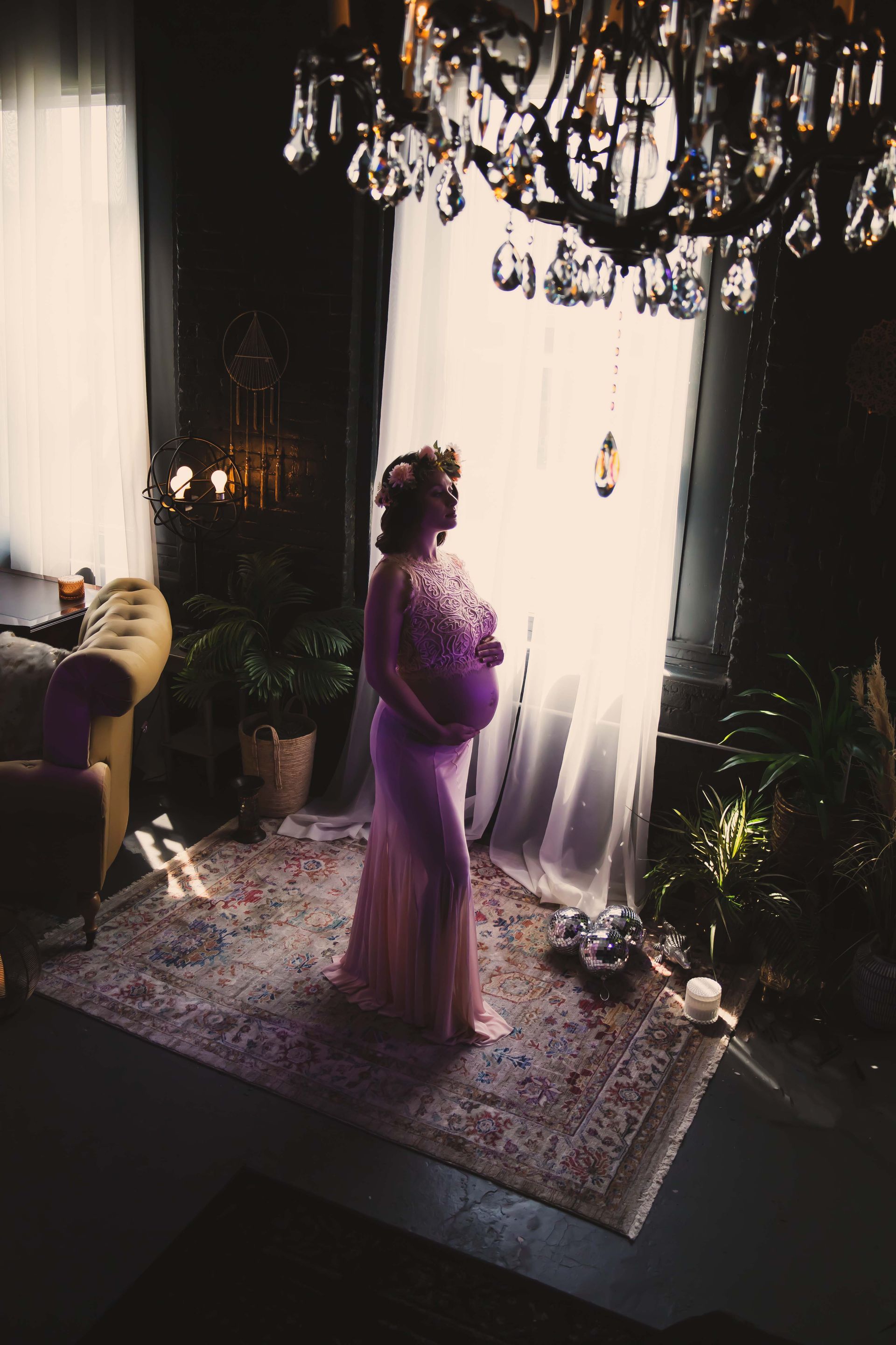 Pregnant woman in a pink gown poses by a bright window in an ornate room, looking pensively outside.