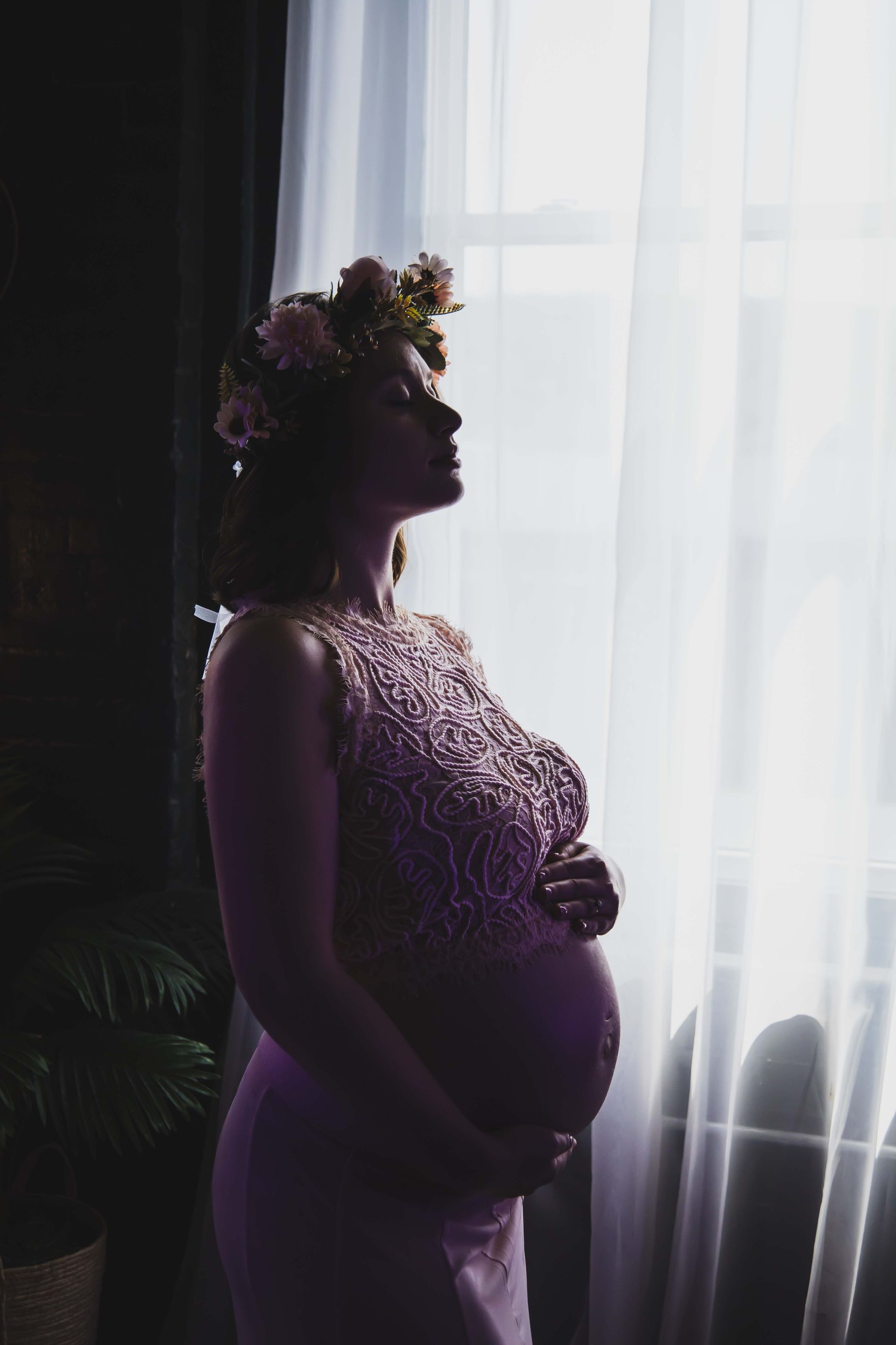 Pregnant woman in a beaded lavender dress and floral crown, gazing out a sunlit window, hands on her belly.