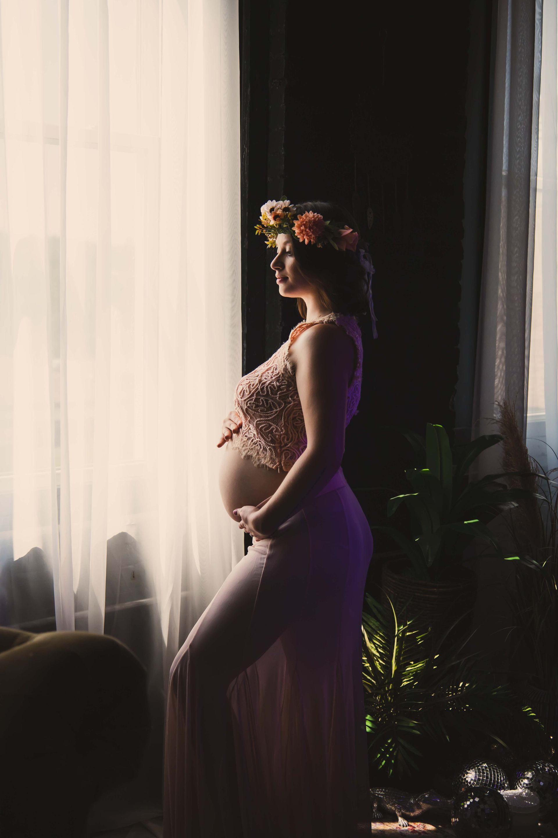 Pregnant woman in a pink dress and flower crown, standing in front of a window and gently touching her belly.