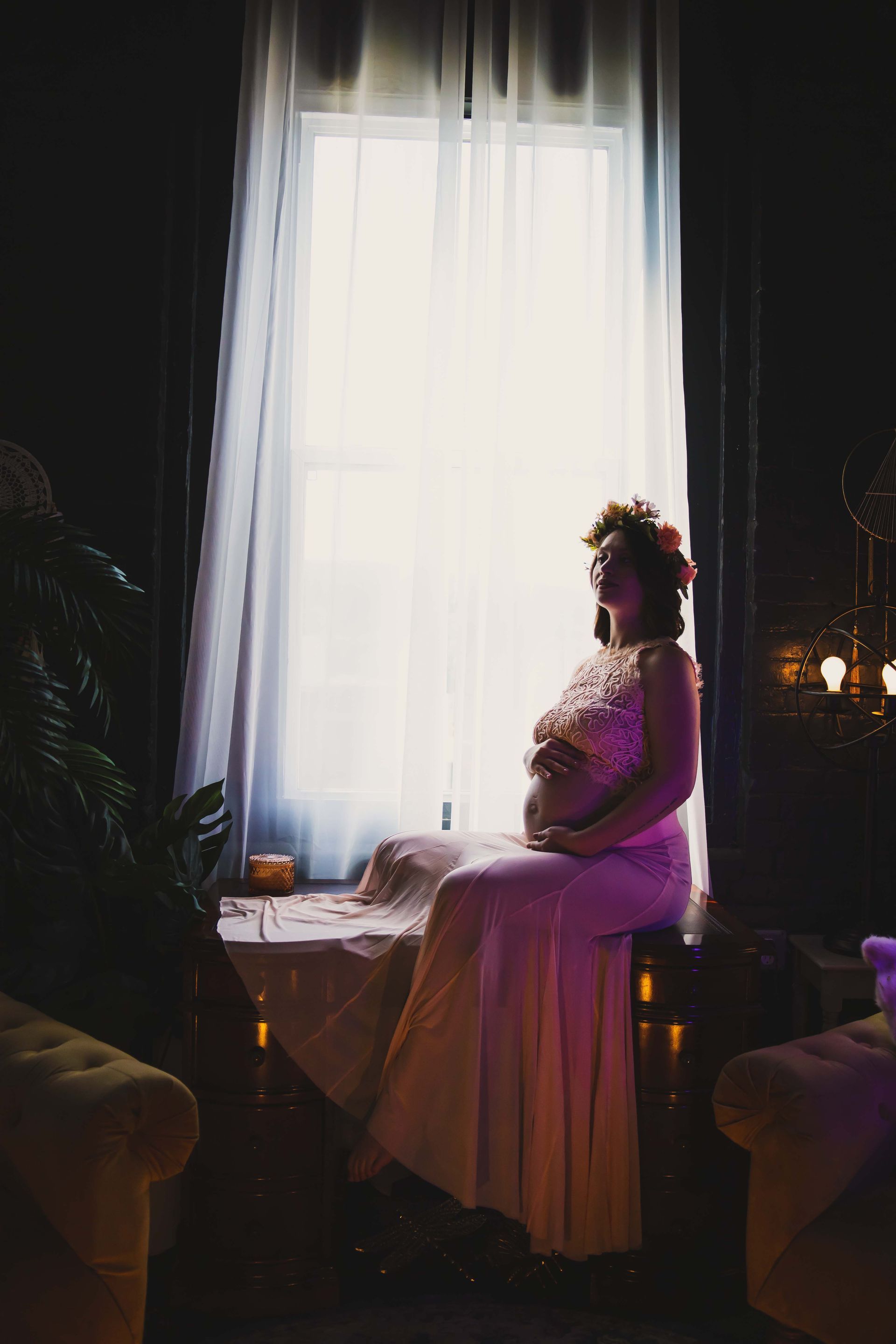 Pregnant woman in a light pink dress and floral crown sits by a sunlit window, holding her belly. Dark room with plants and candles.