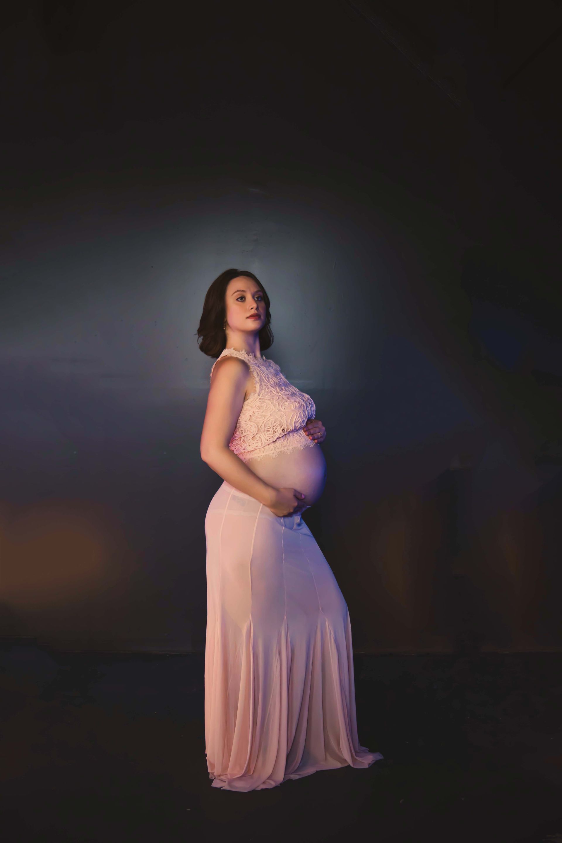 Pregnant woman in a pink dress, cradling her belly. She's facing the camera in a dimly lit studio.
