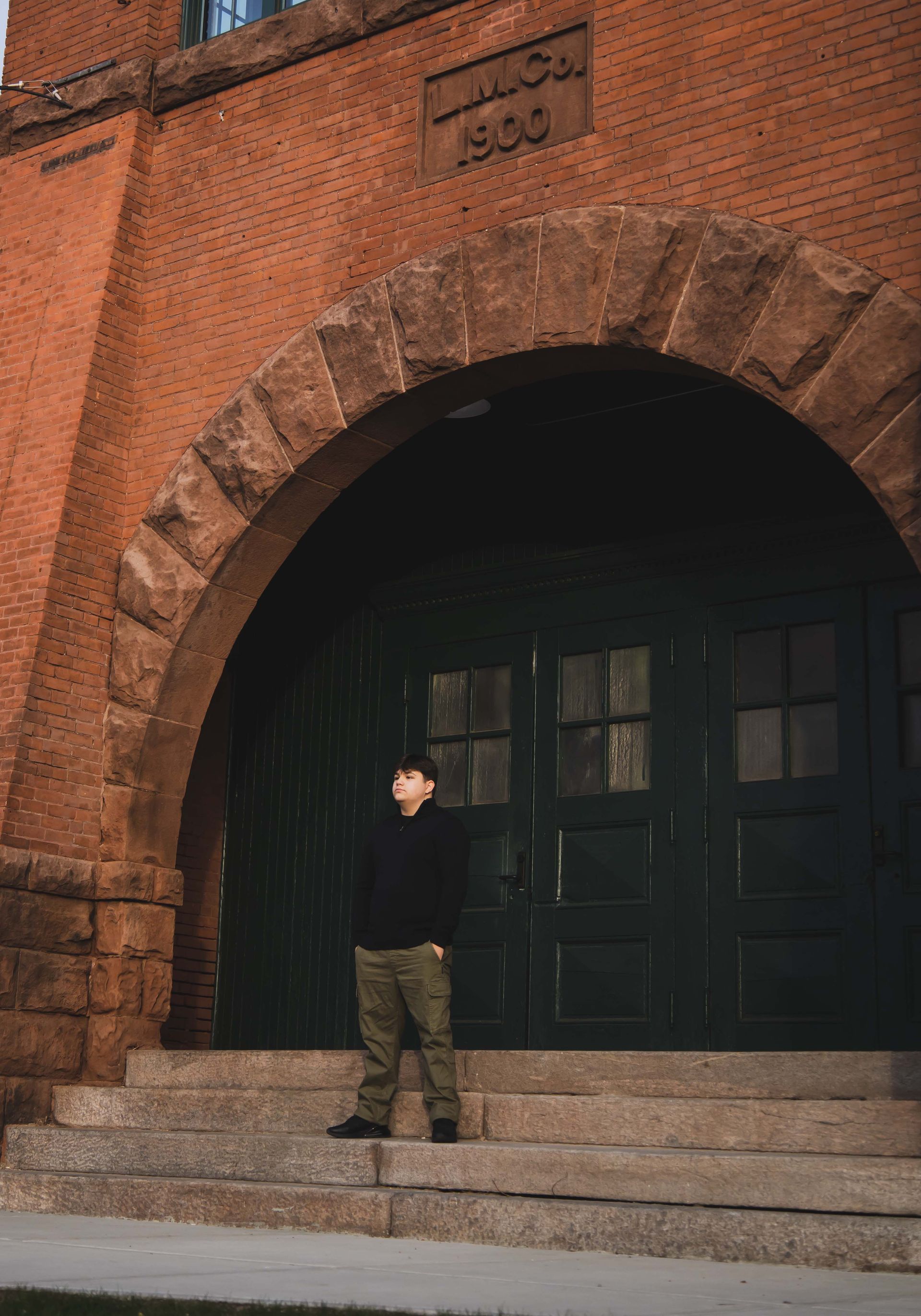 Man standing on steps in front of a brick building with an arched entryway and large green doors. He's wearing dark clothing and khaki pants.