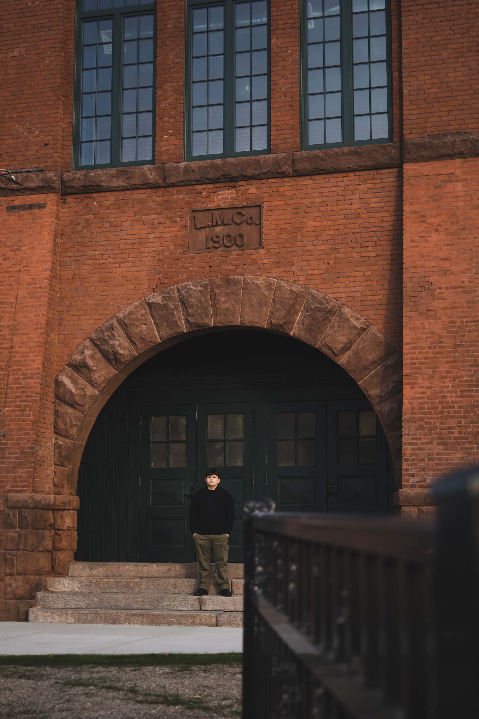 A person stands at the entrance of an old brick building with an arched doorway. The building has green window frames and a 1936 inscription above the entrance.