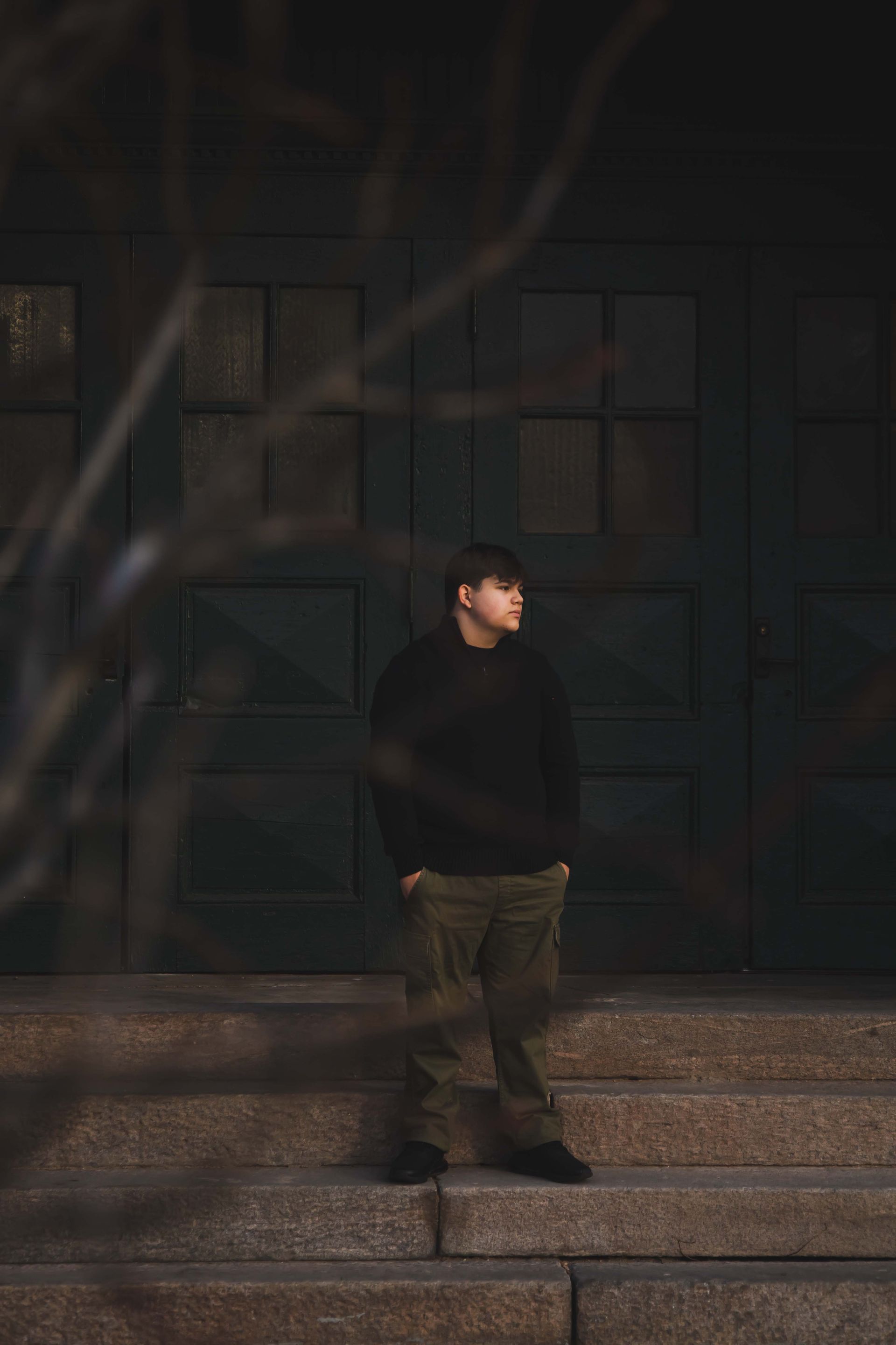 Man in dark clothing stands on stone steps in front of a dark green door, looking off to the side. Branches in the foreground.