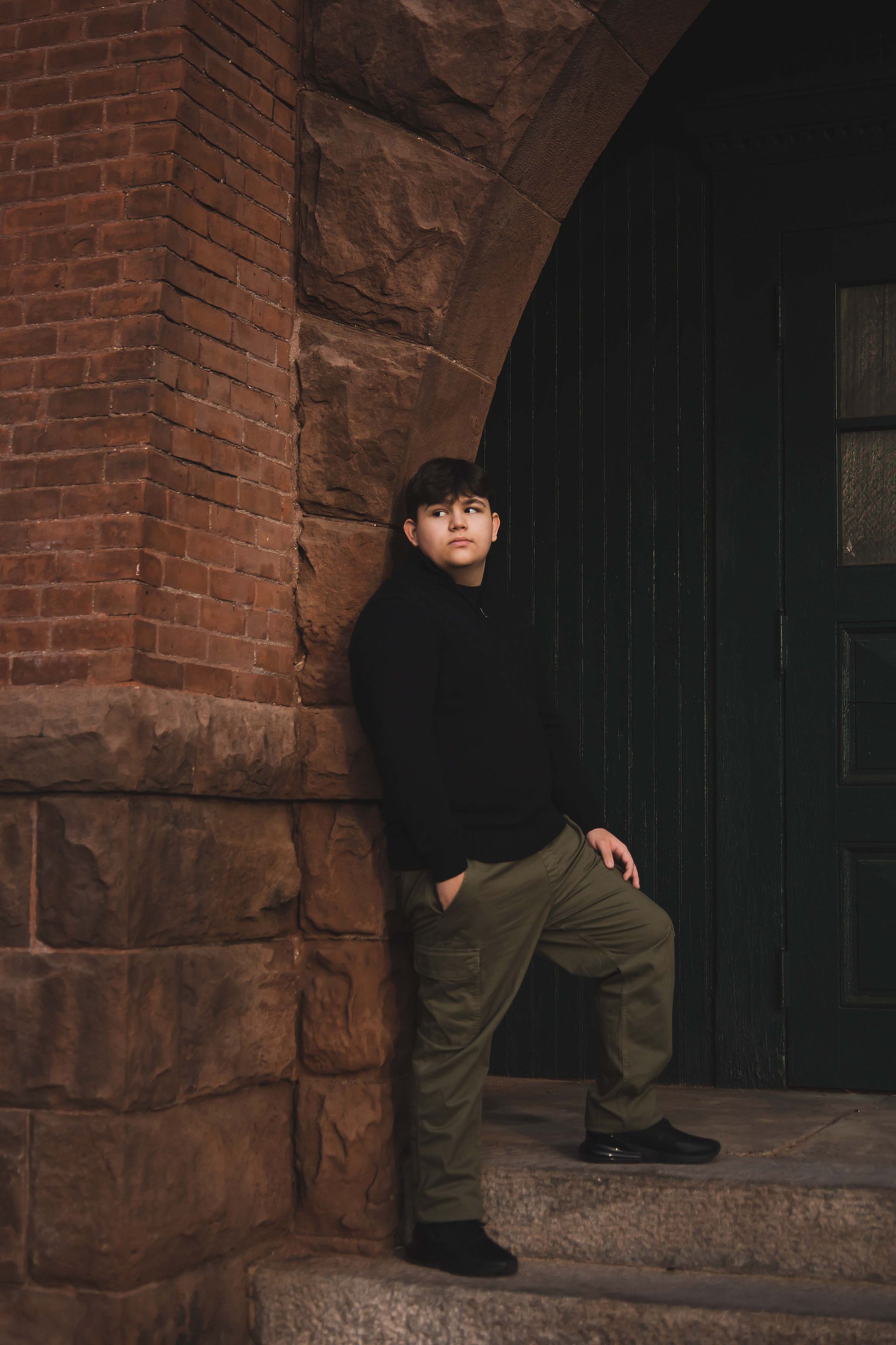 A young man stands on steps next to a brick building. He wears a black hoodie and green pants, looking toward the camera.