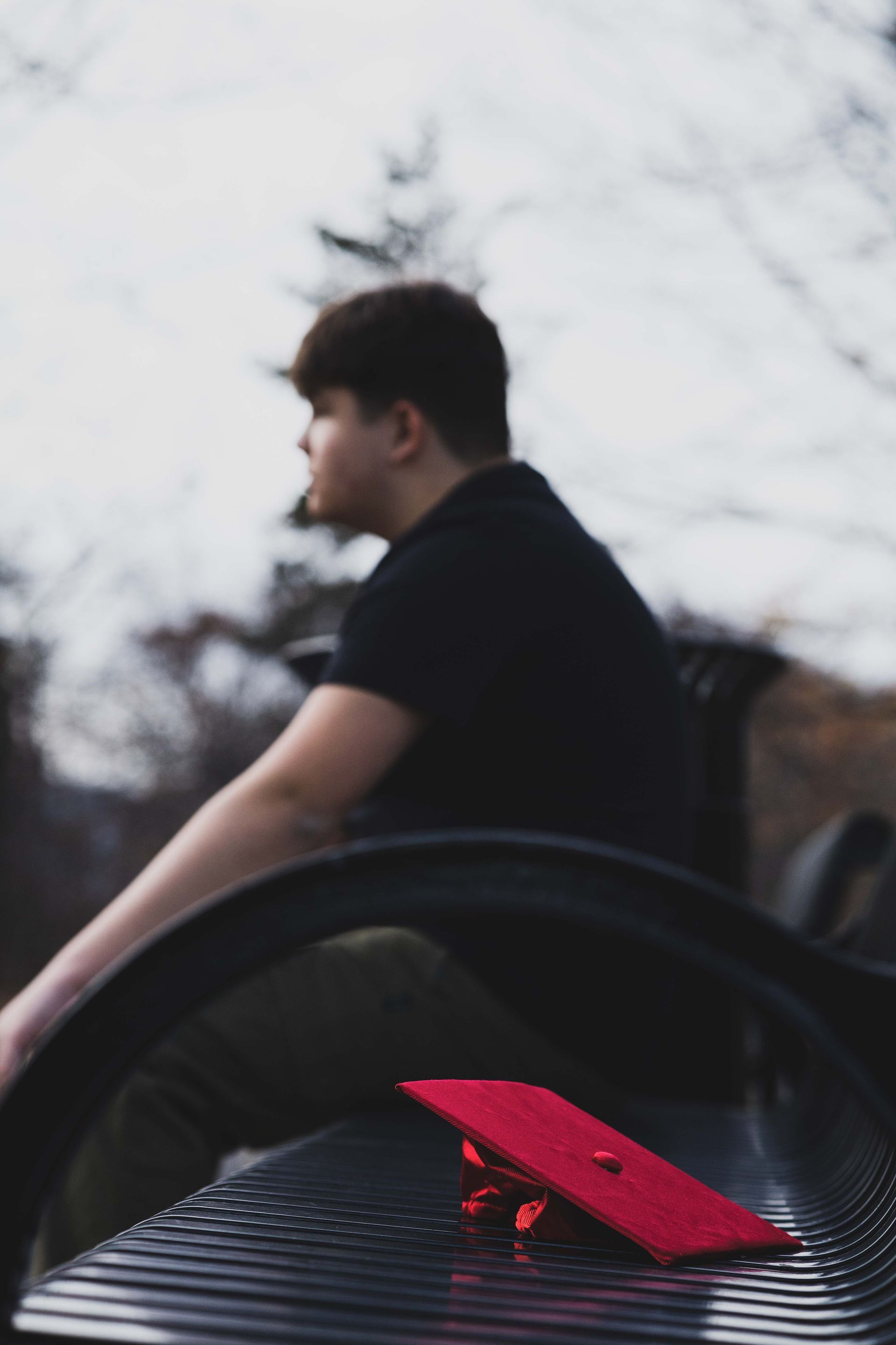 A person in a black shirt sitting on a bench with a red graduation cap on it. Outdoors, blurred background.