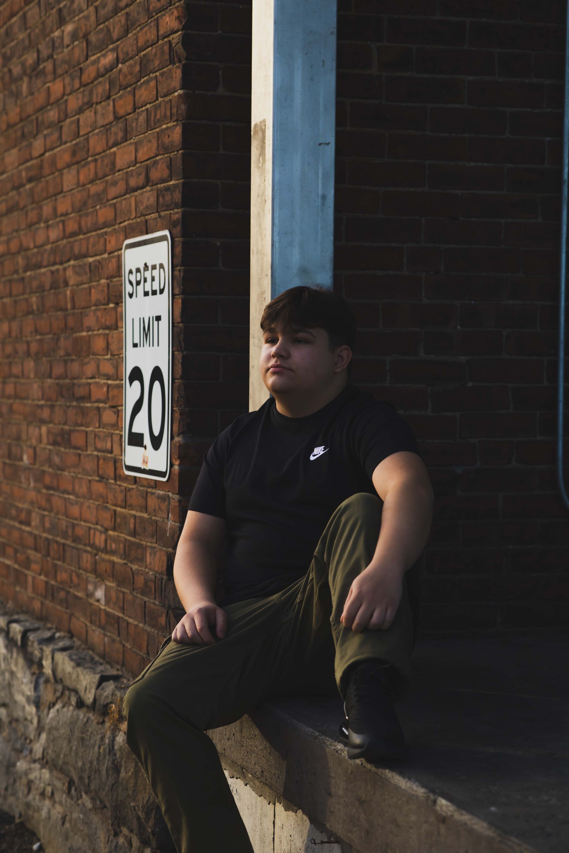 A young person sits on a ledge in front of a brick wall, wearing a black shirt and green pants, with a speed limit sign visible.