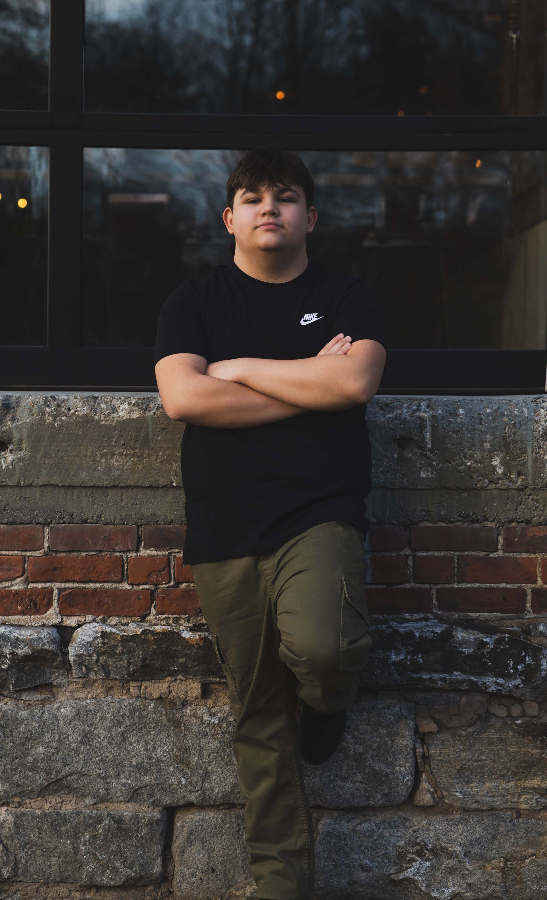 Teenage boy in a black shirt and green pants leaning against a brick wall, arms crossed, with a serious expression.