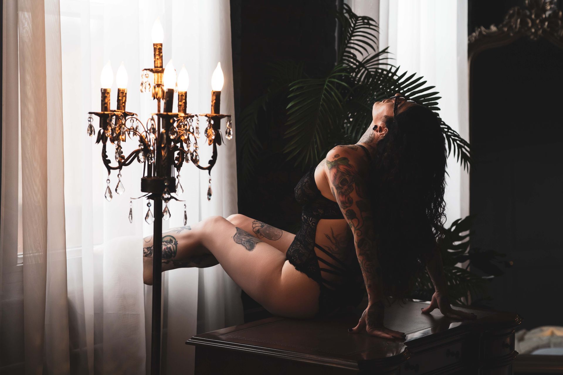 Woman with tattoos in lingerie poses by a window, lit by a candelabra.  Dark room with a green plant and sheer curtains.