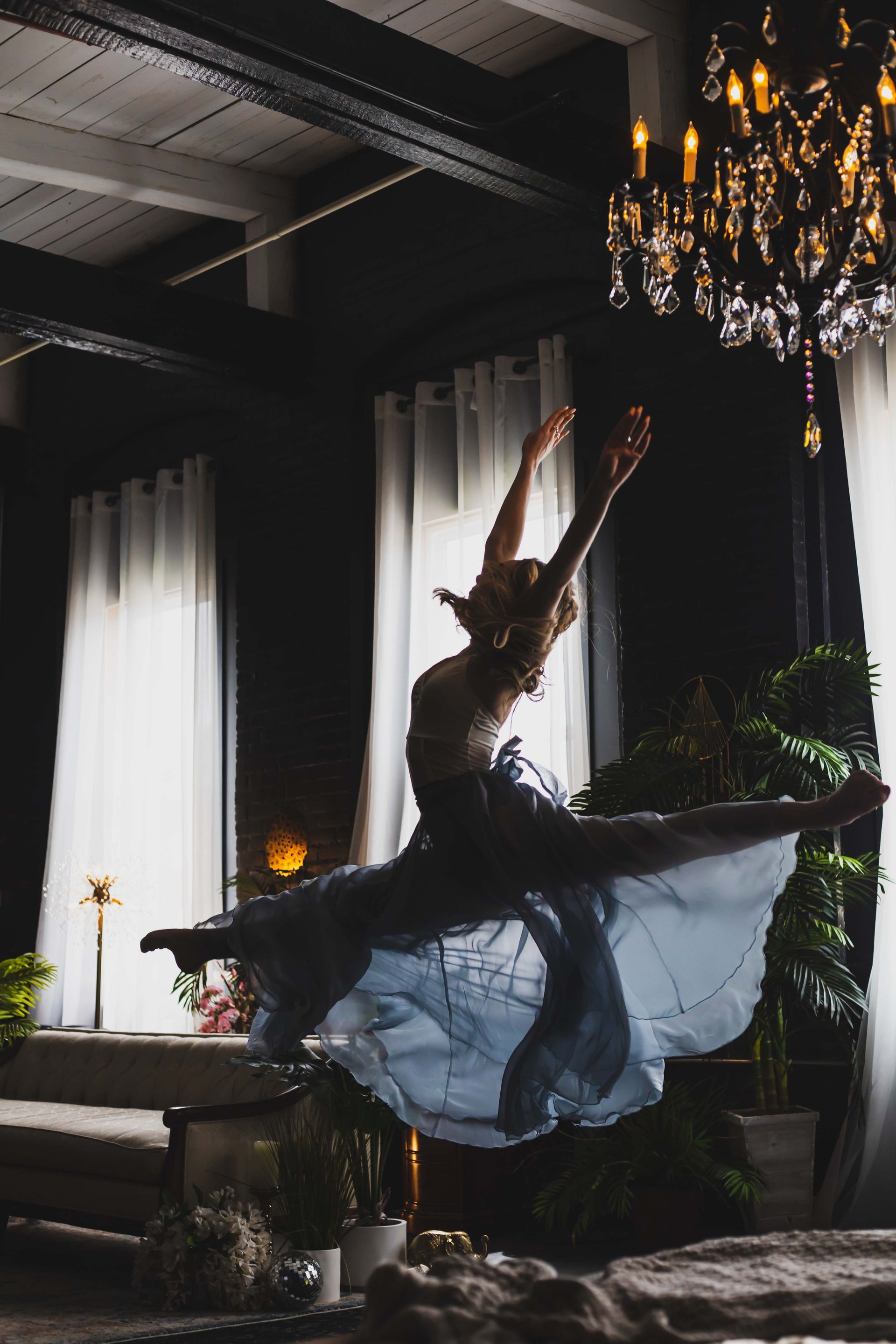 A ballerina leaps gracefully in a dimly lit room, arms extended, wearing a flowing blue dress, with a chandelier overhead.