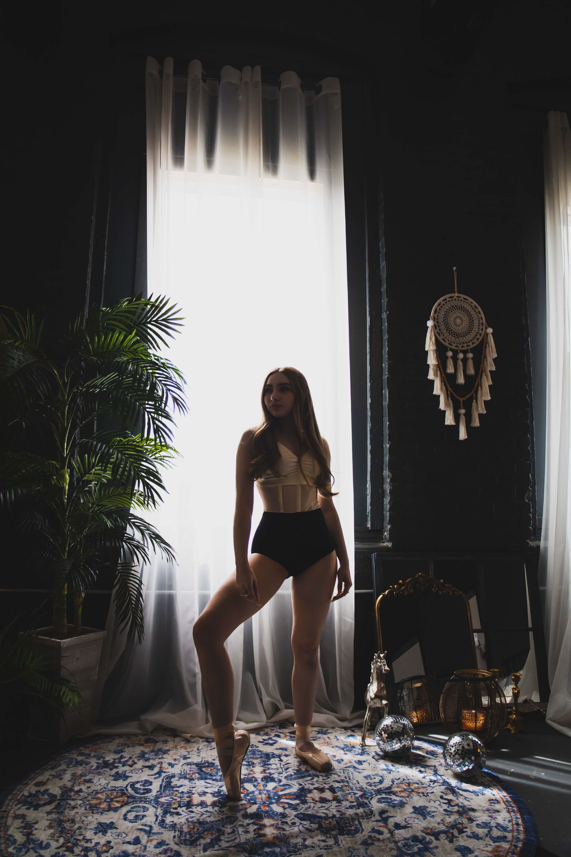 A ballerina in lingerie poses in front of a bright window. She stands en pointe on a patterned rug in a dark-walled room.