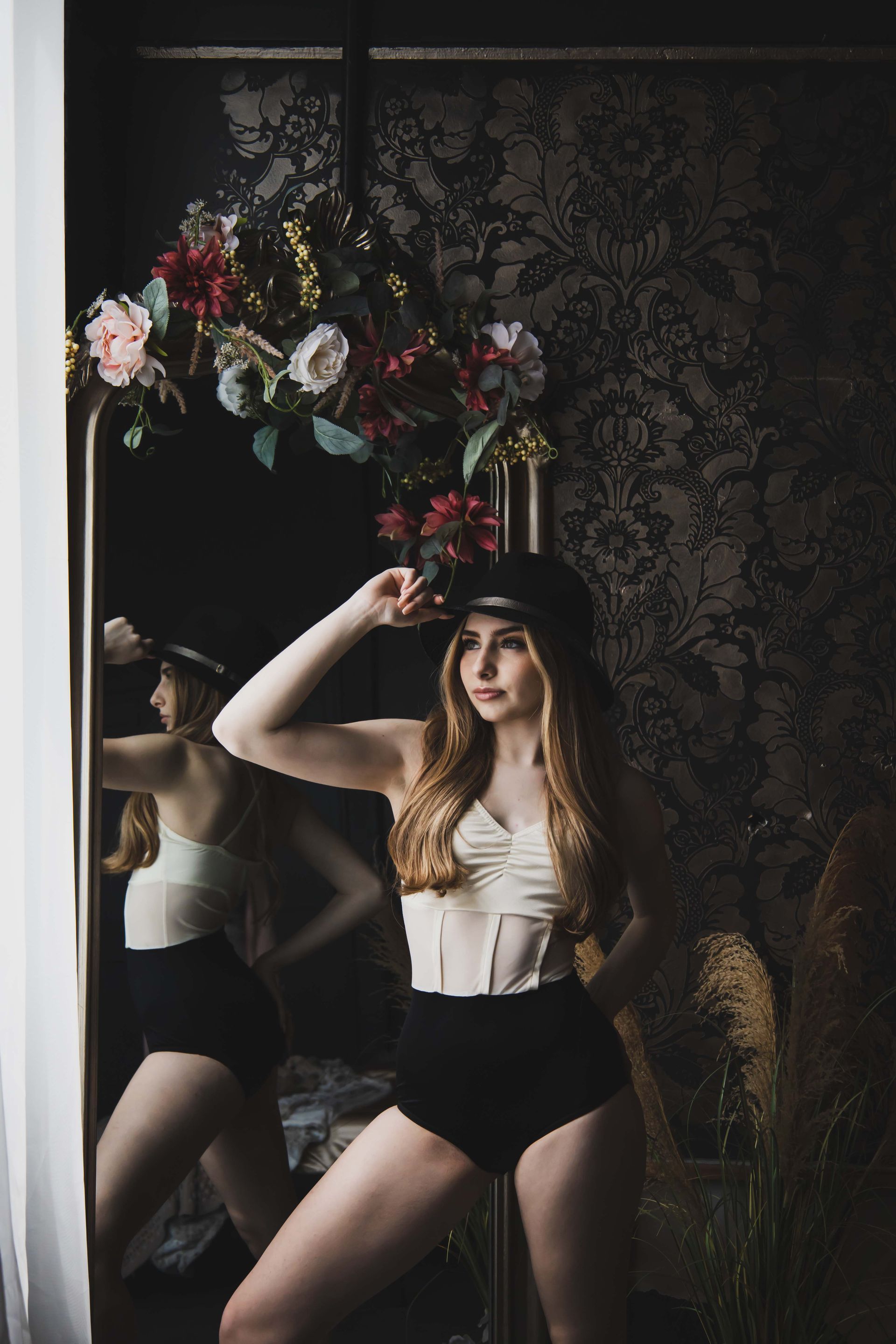 Woman in a black cap and high-waisted shorts poses in front of a mirror, holding her hat. Dark room with floral decoration.