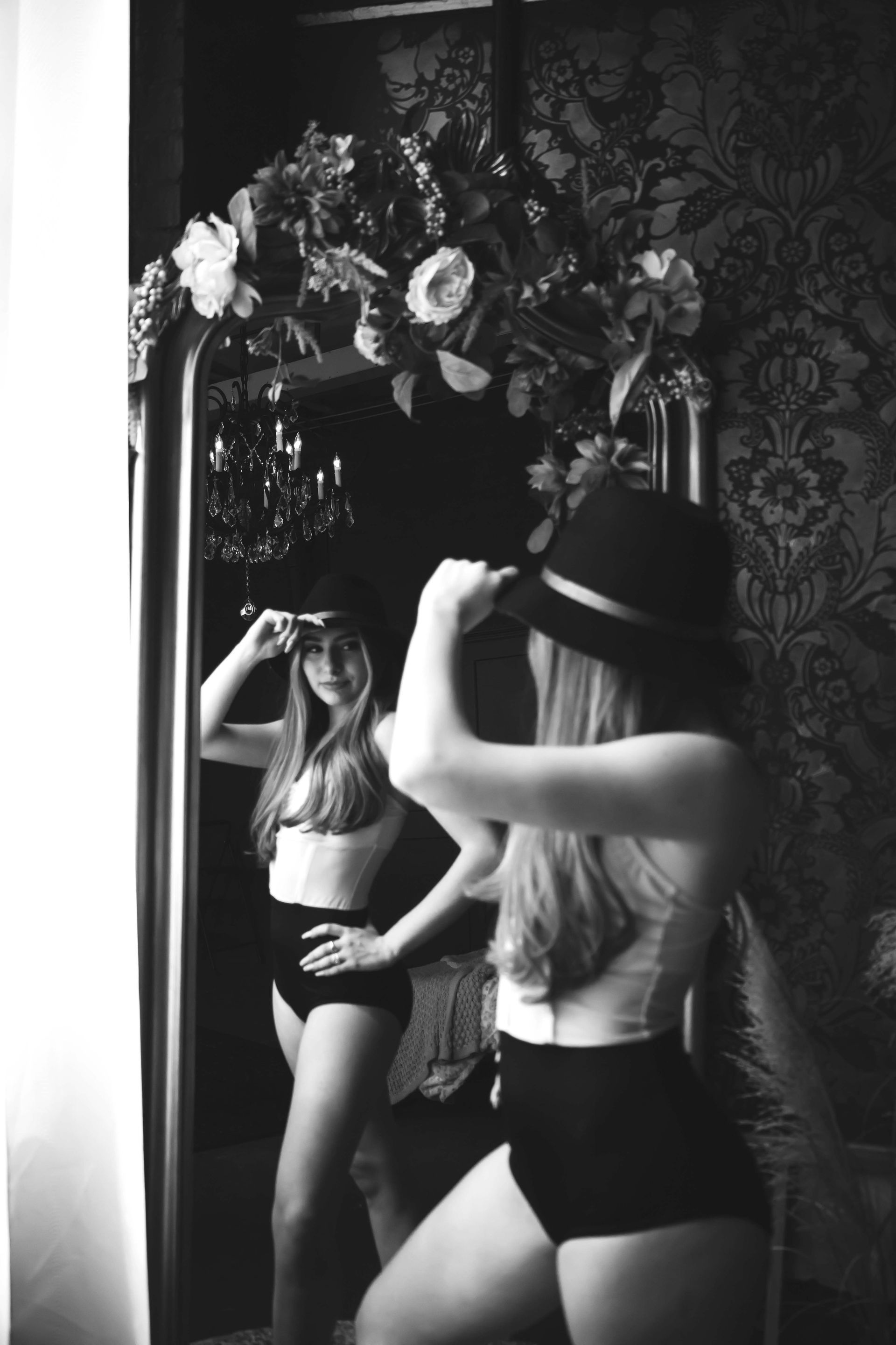 Woman in black shorts and white top, reflected in a large mirror. She is adjusting a black hat in a room with floral wallpaper.