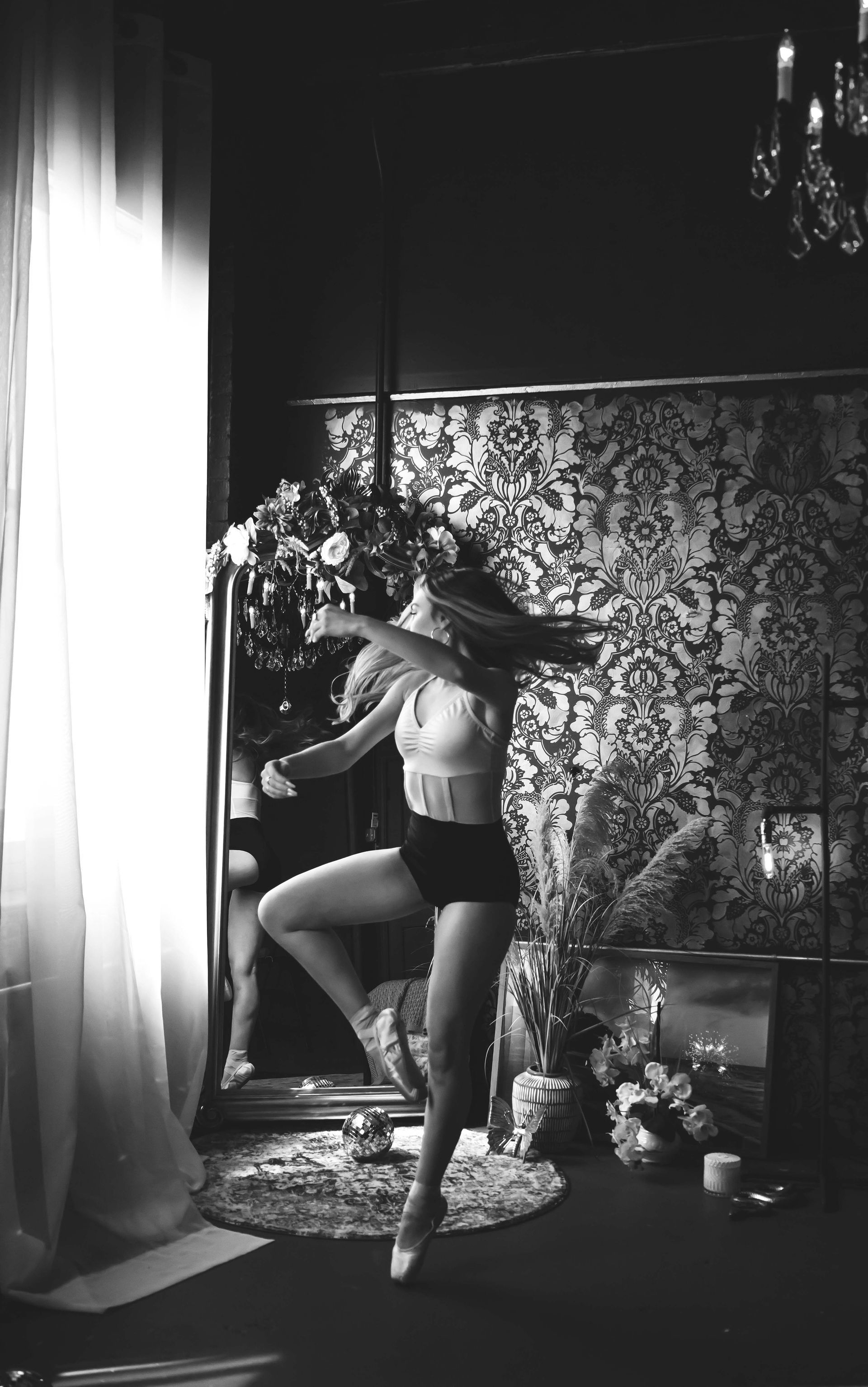 A dancer in lingerie practices on a pole in a room with a decorative backdrop, flowers, and natural light.