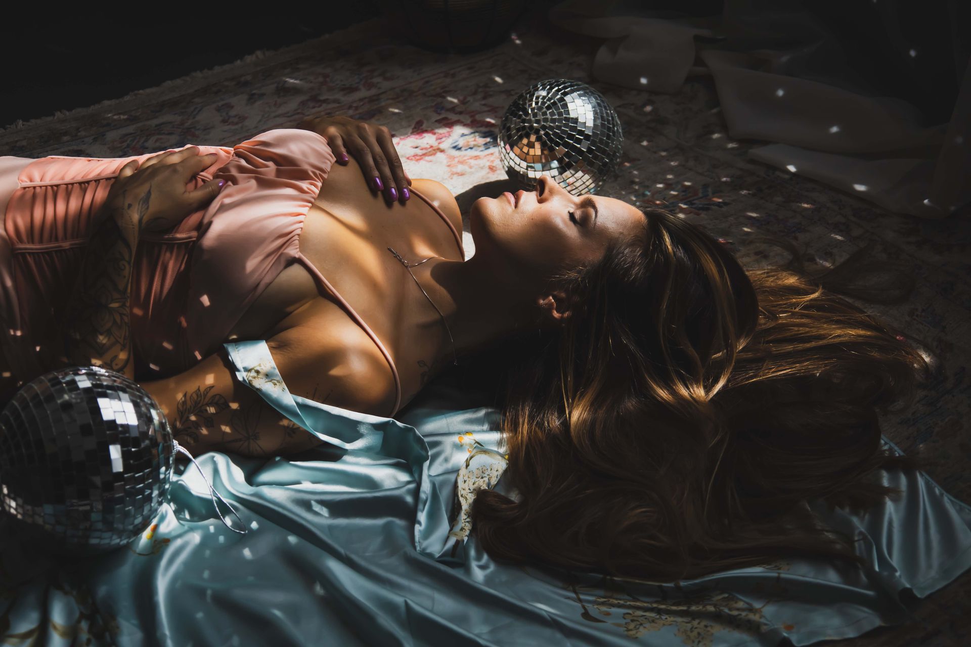 Woman reclining on bed with a pink dress, lit by light from disco balls.