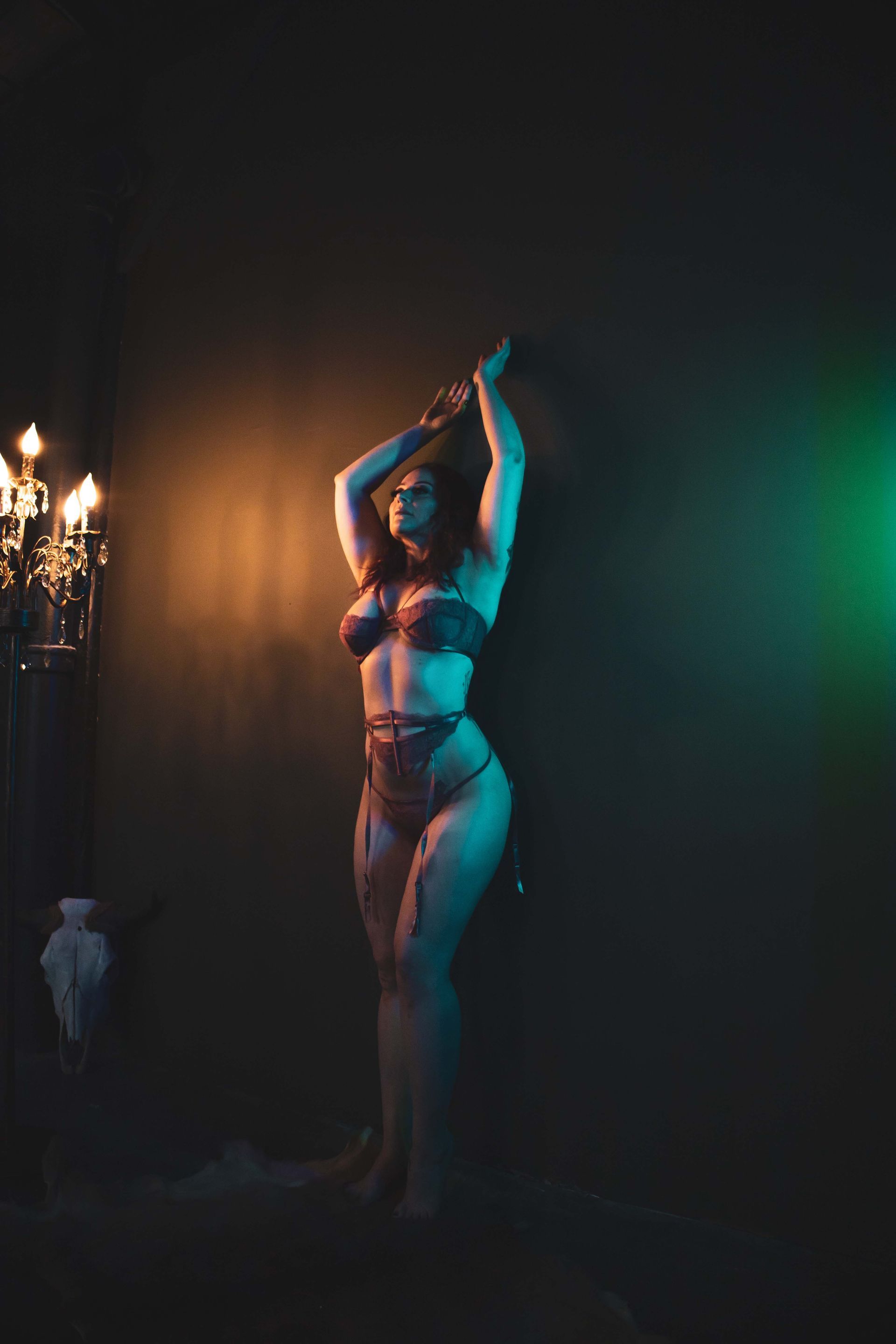 Woman in red lingerie poses against a dark wall with chained arms, under orange and green lighting. A chandelier is visible to the left.