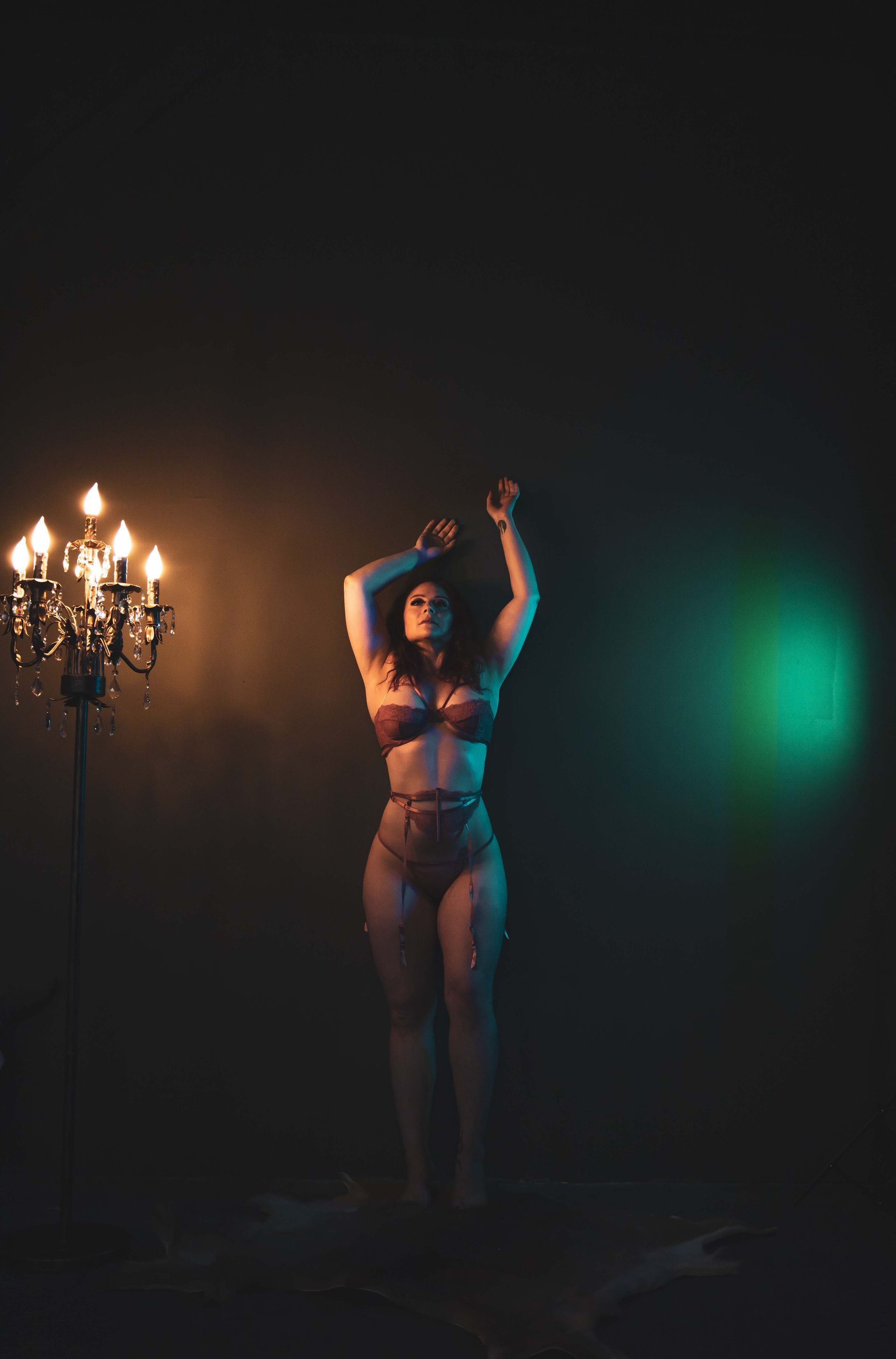 Woman in lingerie, arms raised, lit by a chandelier and colored spotlights against a dark backdrop.