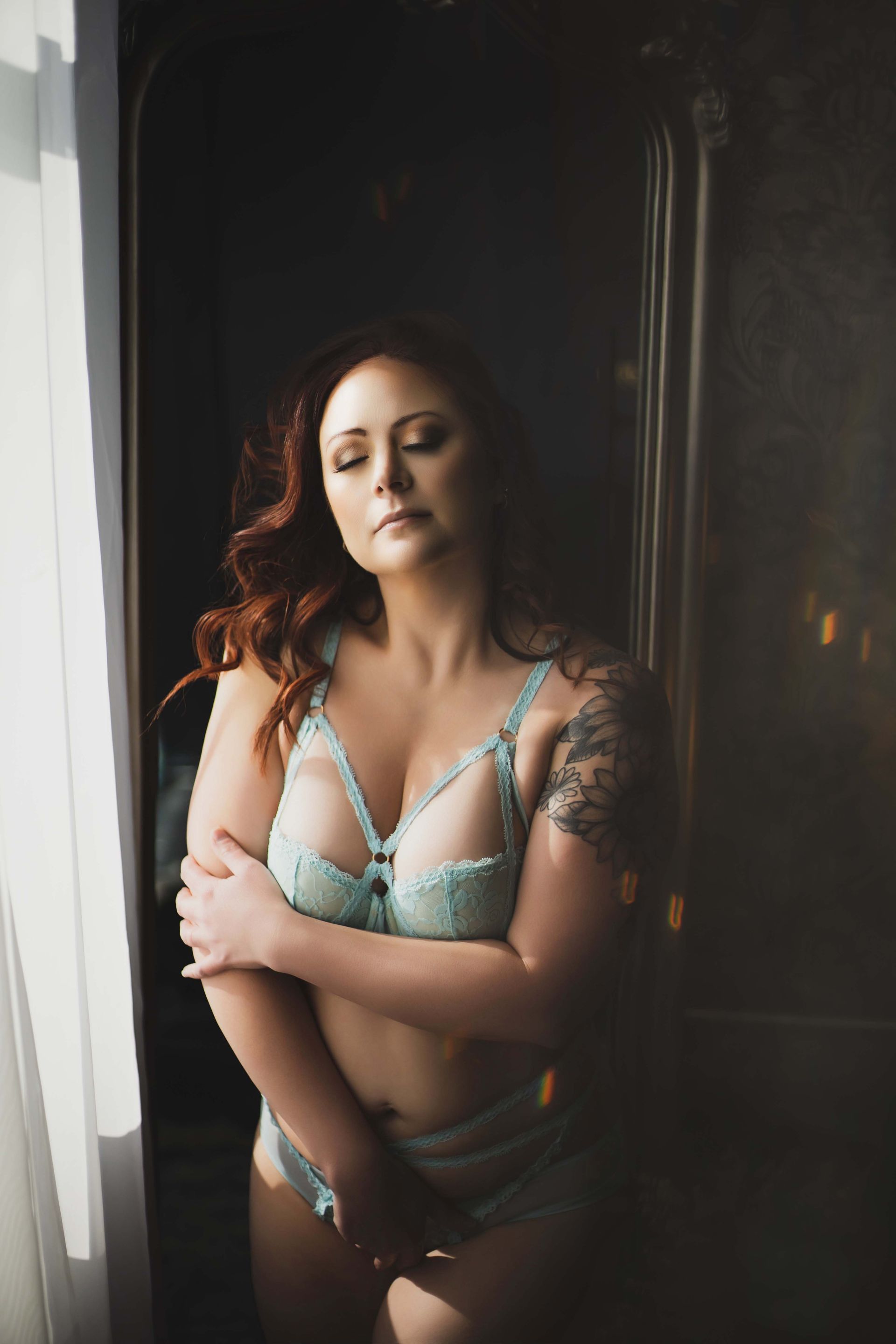 Woman with red hair wearing light blue lingerie, arms crossed, stands in front of a window and ornate dark door.