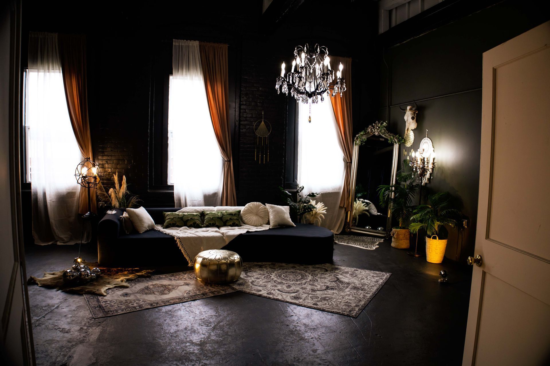 Dim living room with white curtains, a dark sofa, chandelier, rug, and potted plants by a window