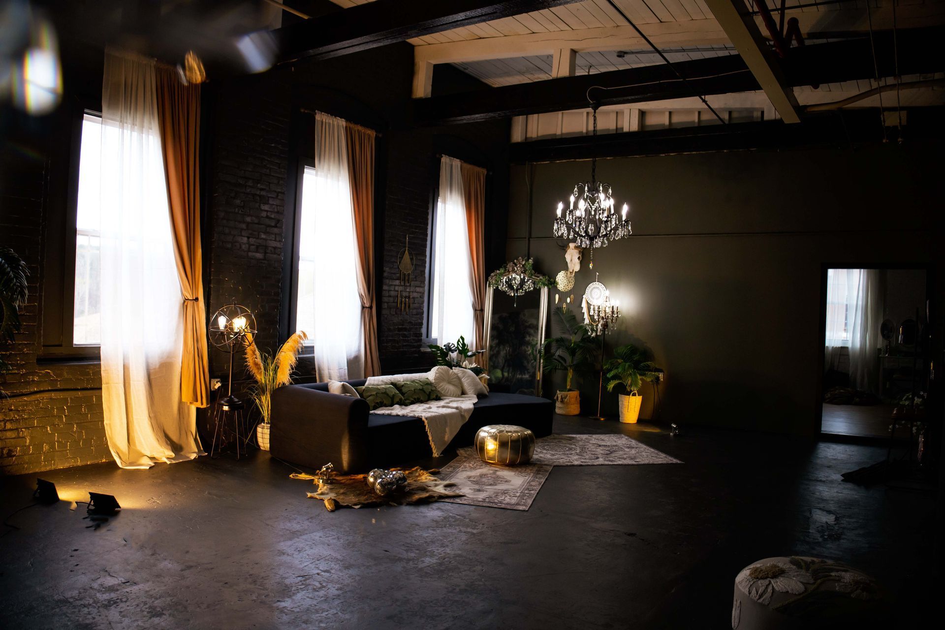 Elegant dark lounge with tall lit windows, a green sofa, chandeliers, and potted plants near a large mirror