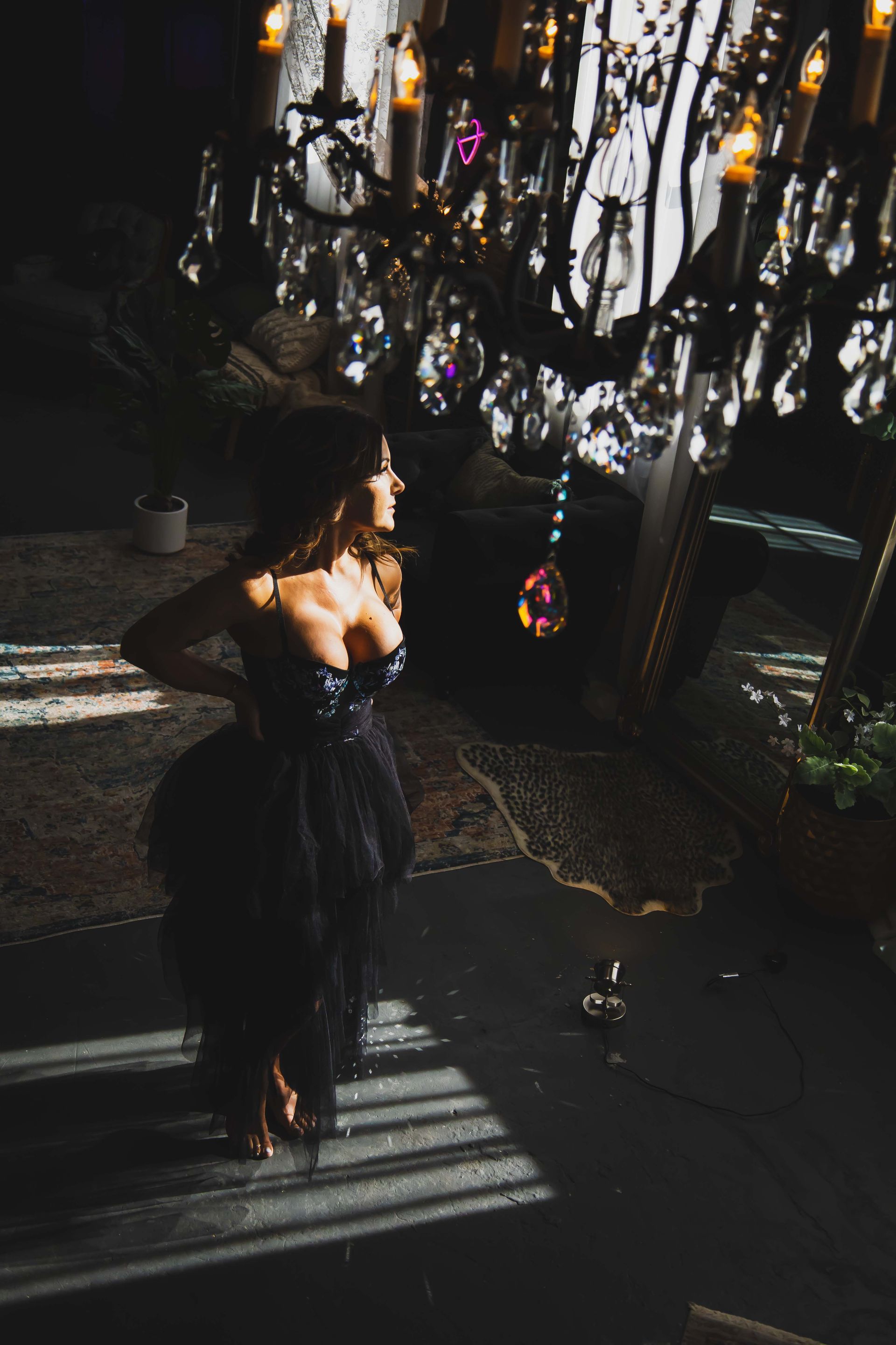 Woman in a black gown stands under a chandelier. Dramatic lighting casts shadows.