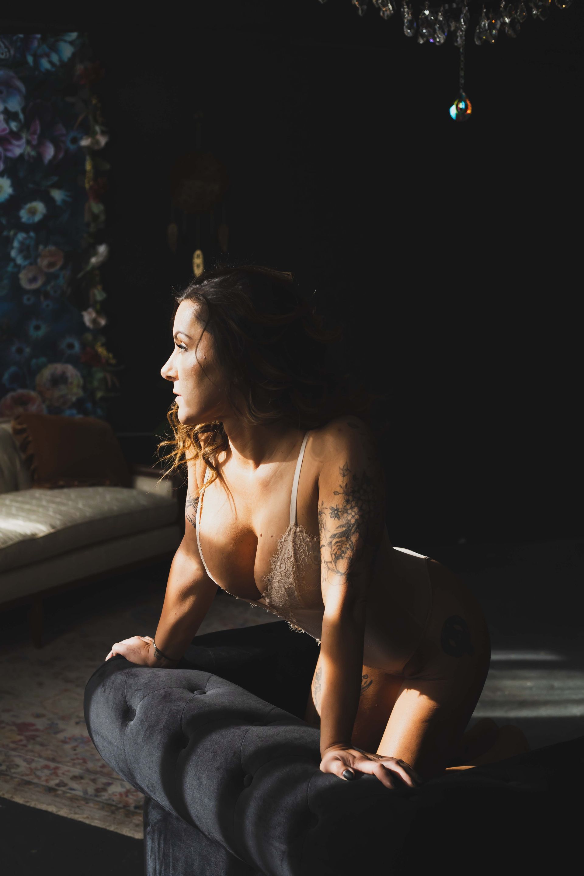 Woman in lingerie, looking out a window, lit by sunlight. Leaning on a dark couch in a dimly lit room.