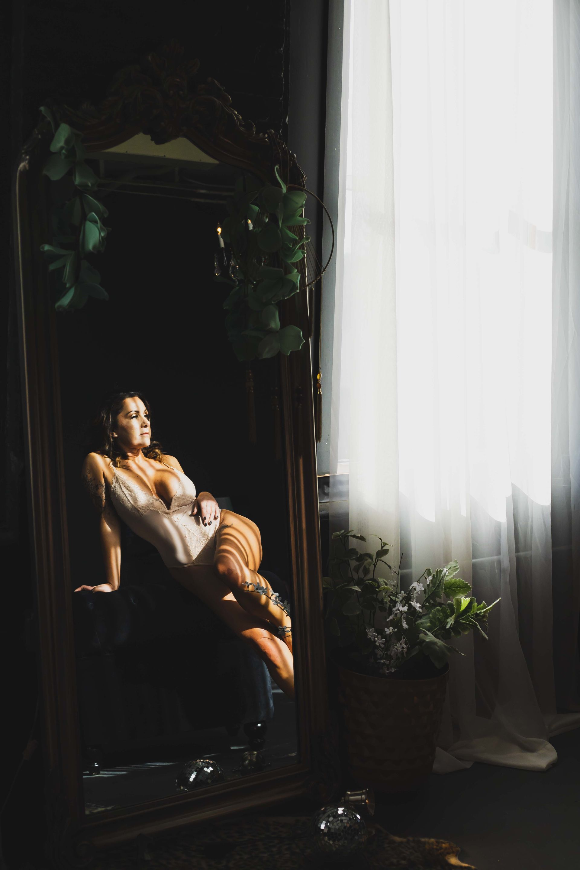 Woman in lingerie poses near a large mirror in a dimly lit room, sunlight streaming in from a window.