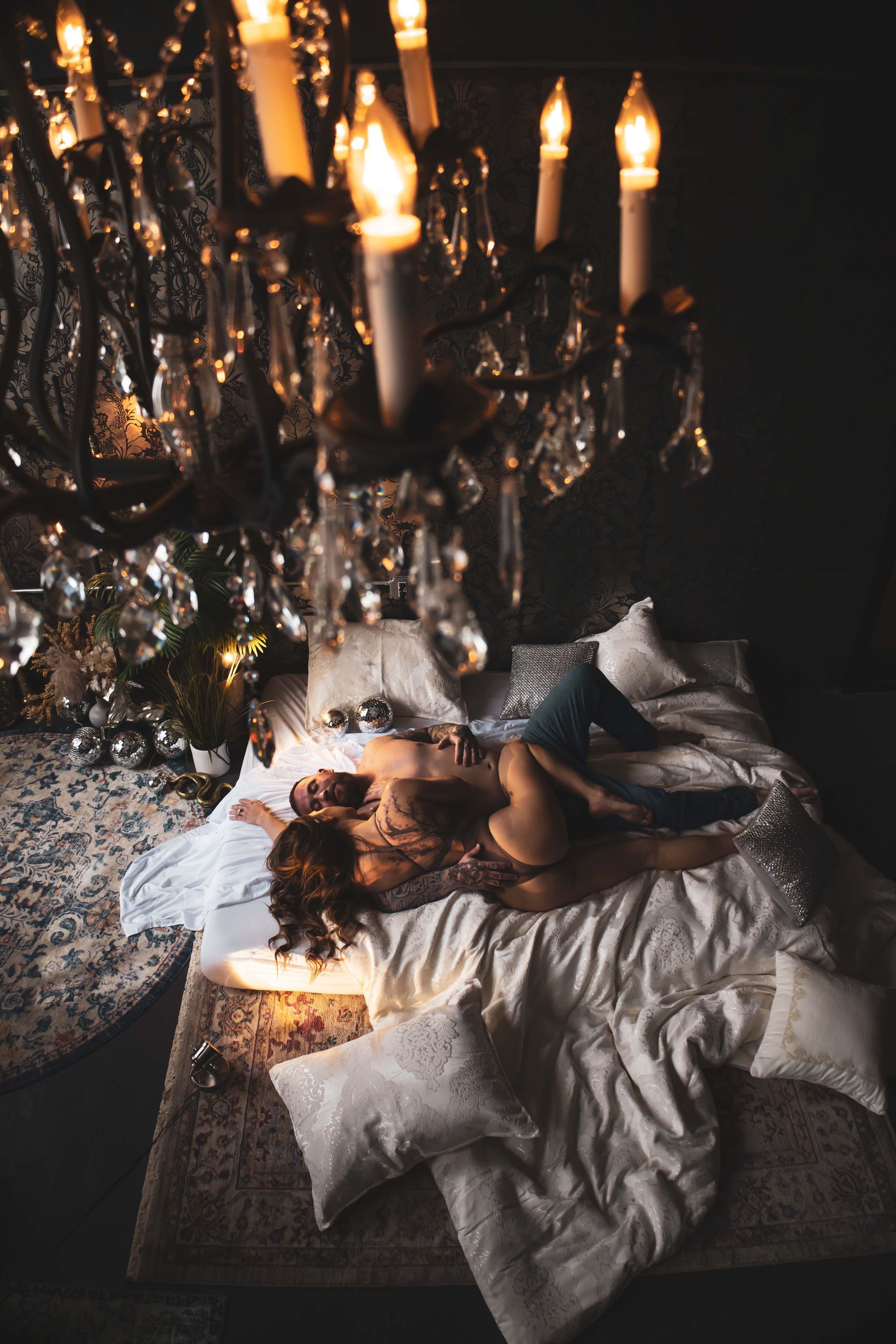 Woman lies on a bed beneath a sparkling chandelier. The scene is dimly lit, with soft lighting and a glamorous feel.