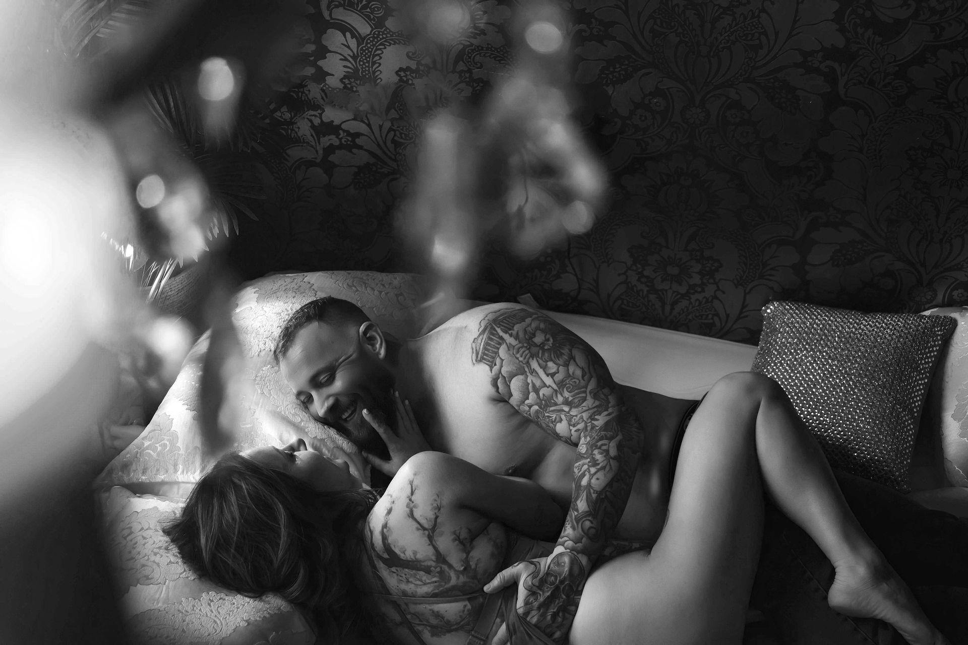 A man and woman embrace intimately on a couch. They are shirtless and covered in tattoos, with the man leaning over the woman.